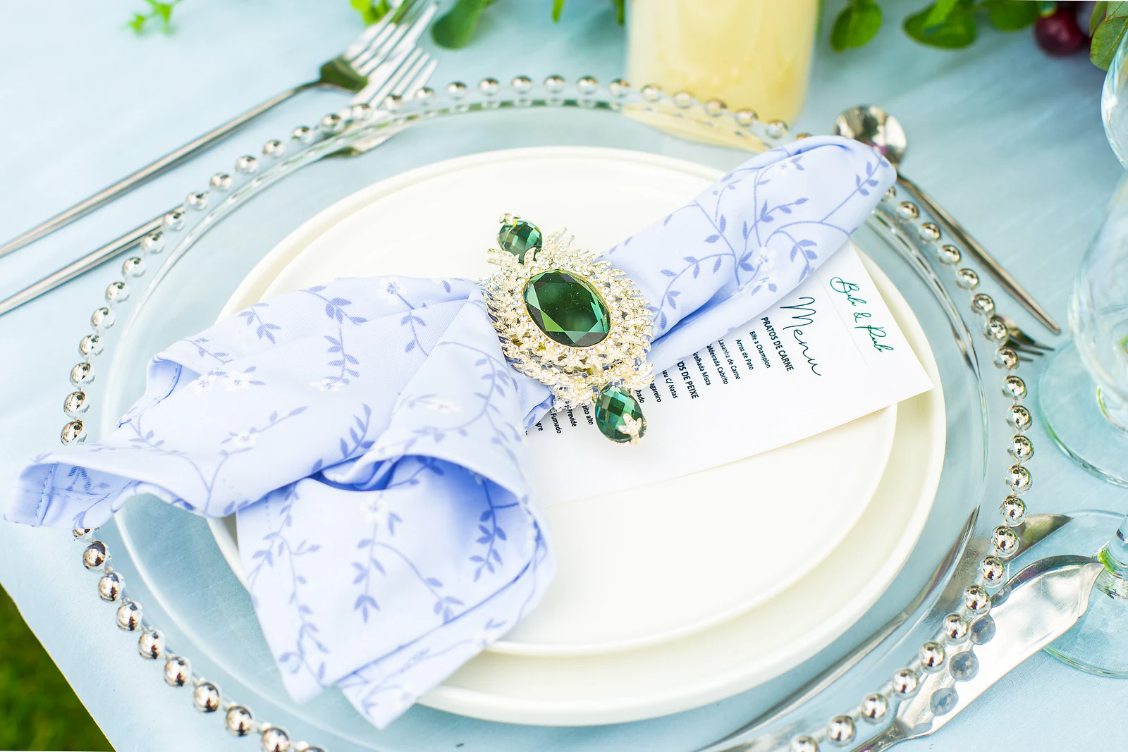 A plate on a beaded glass charger, with a lilac patterned napkin held by a ring set with a green stone and a menu card on top.