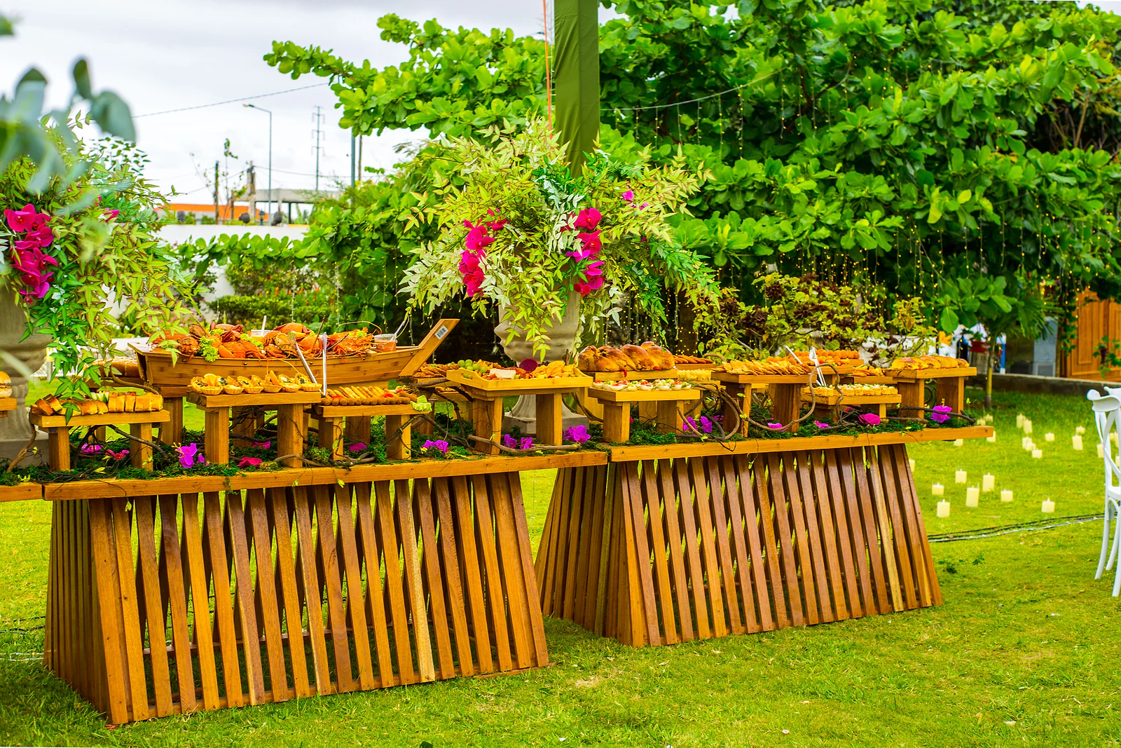 A buffet on slatted timber tables with wooden stands, fuchsia flowers and trailing foliage, set on the lawn.