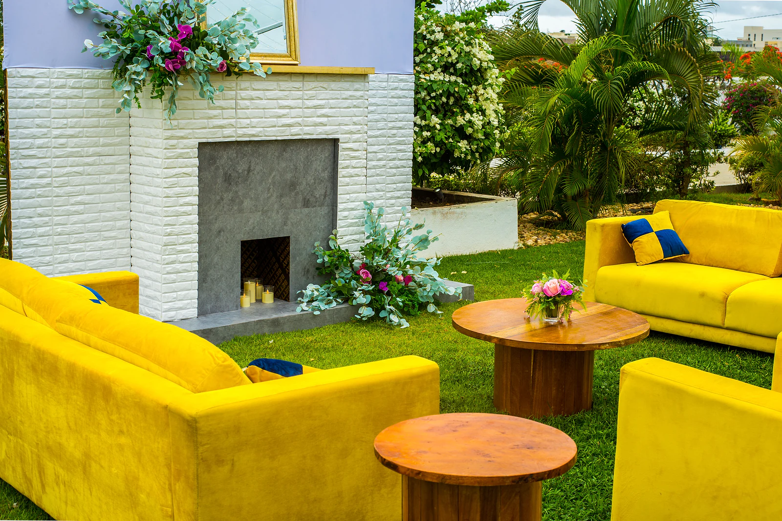 Yellow velvet sofas and round wooden tables facing a white brick fireplace dressed with flowers.