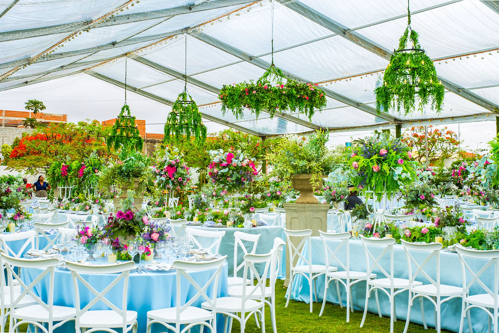 The glass marquee seen from inside, with foliage chandeliers hanging above pale blue tables, white chairs and rose arrangements.