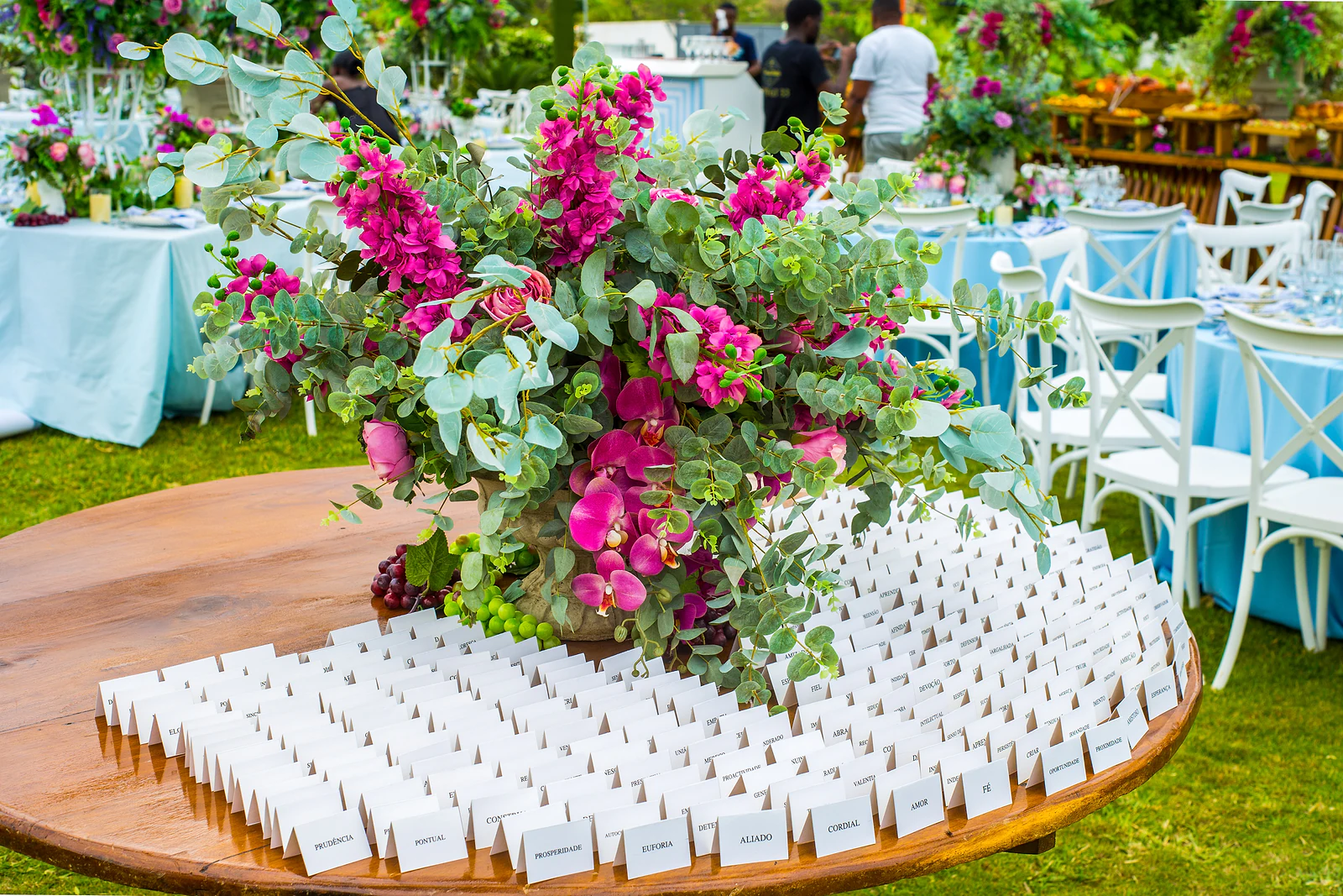 A round wooden table covered in white place cards, with a large arrangement of fuchsia flowers and eucalyptus.