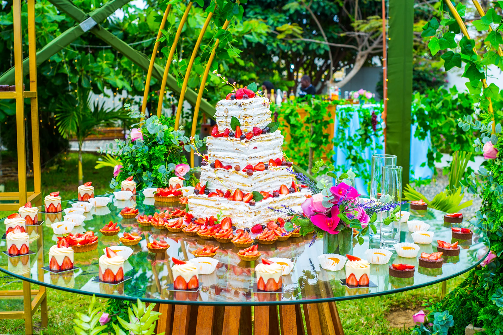 A multi-tiered strawberry cake on a glass table surrounded by sweets, between golden arches and foliage.