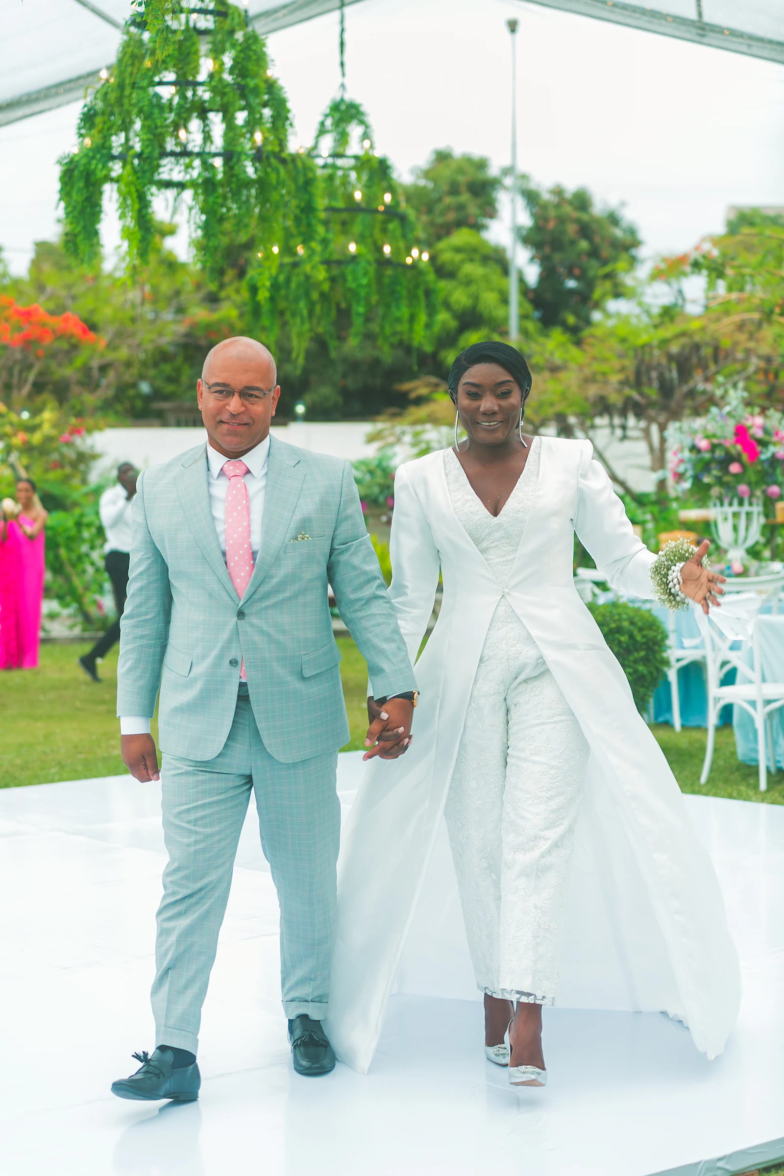 The couple walking hand in hand across the white dance floor, with a foliage chandelier overhead and tables behind.
