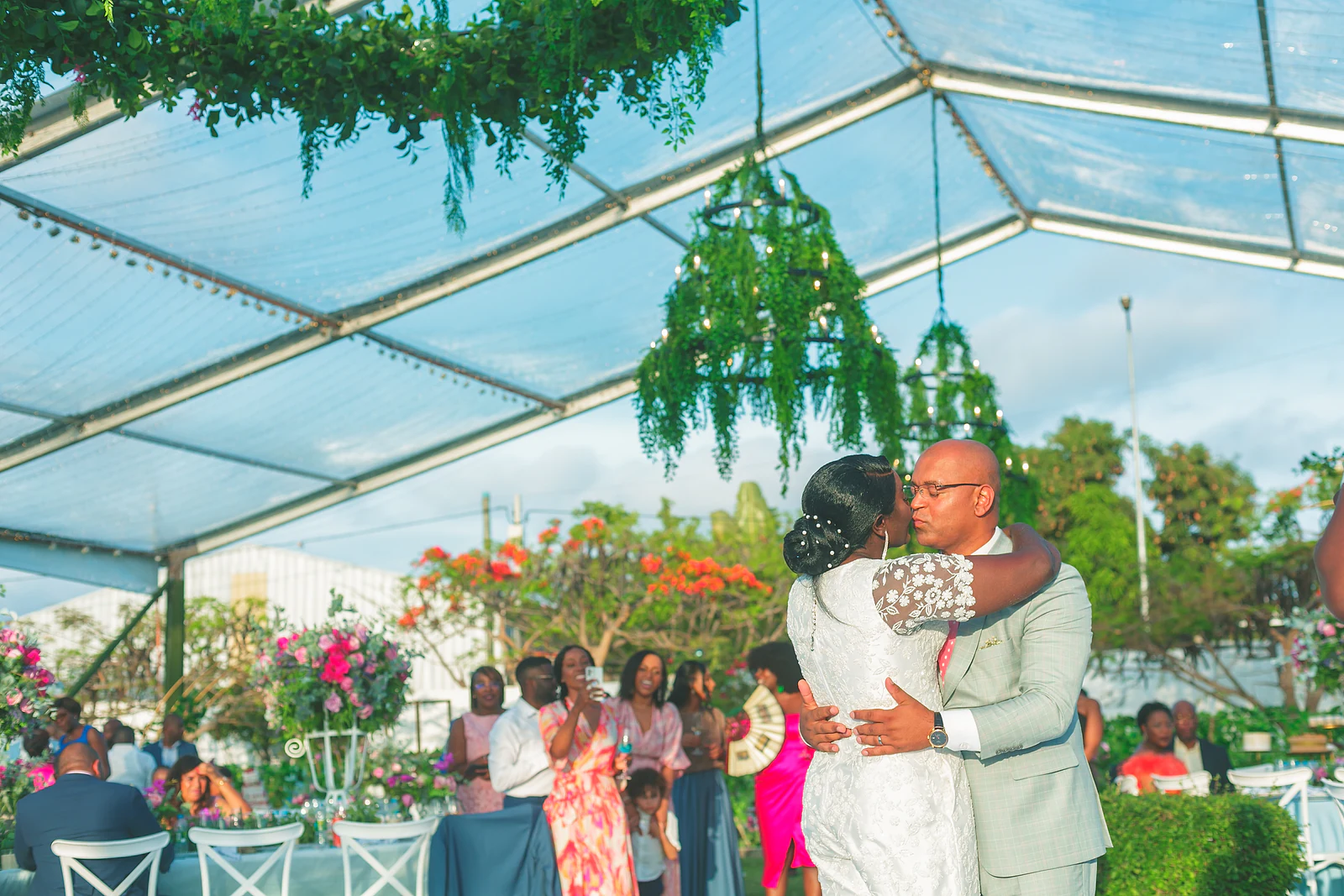 The couple embrace on the white dance floor beneath the marquee's foliage chandeliers, with guests behind them.