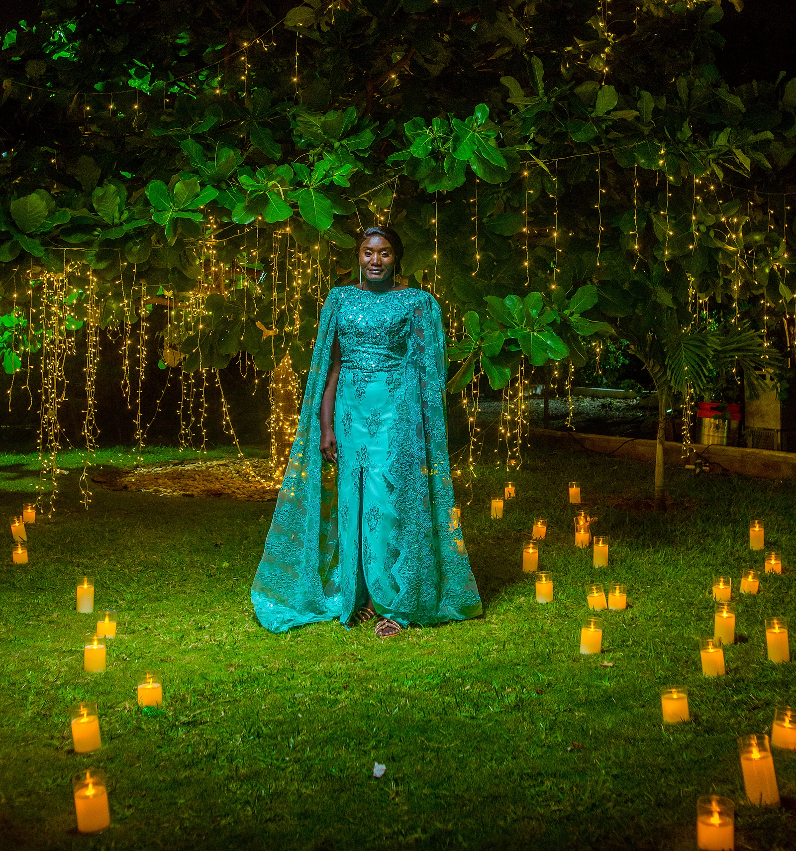A woman in a turquoise cape dress standing beneath a tree hung with curtains of lights, with candles scattered across the lawn at nightfall.