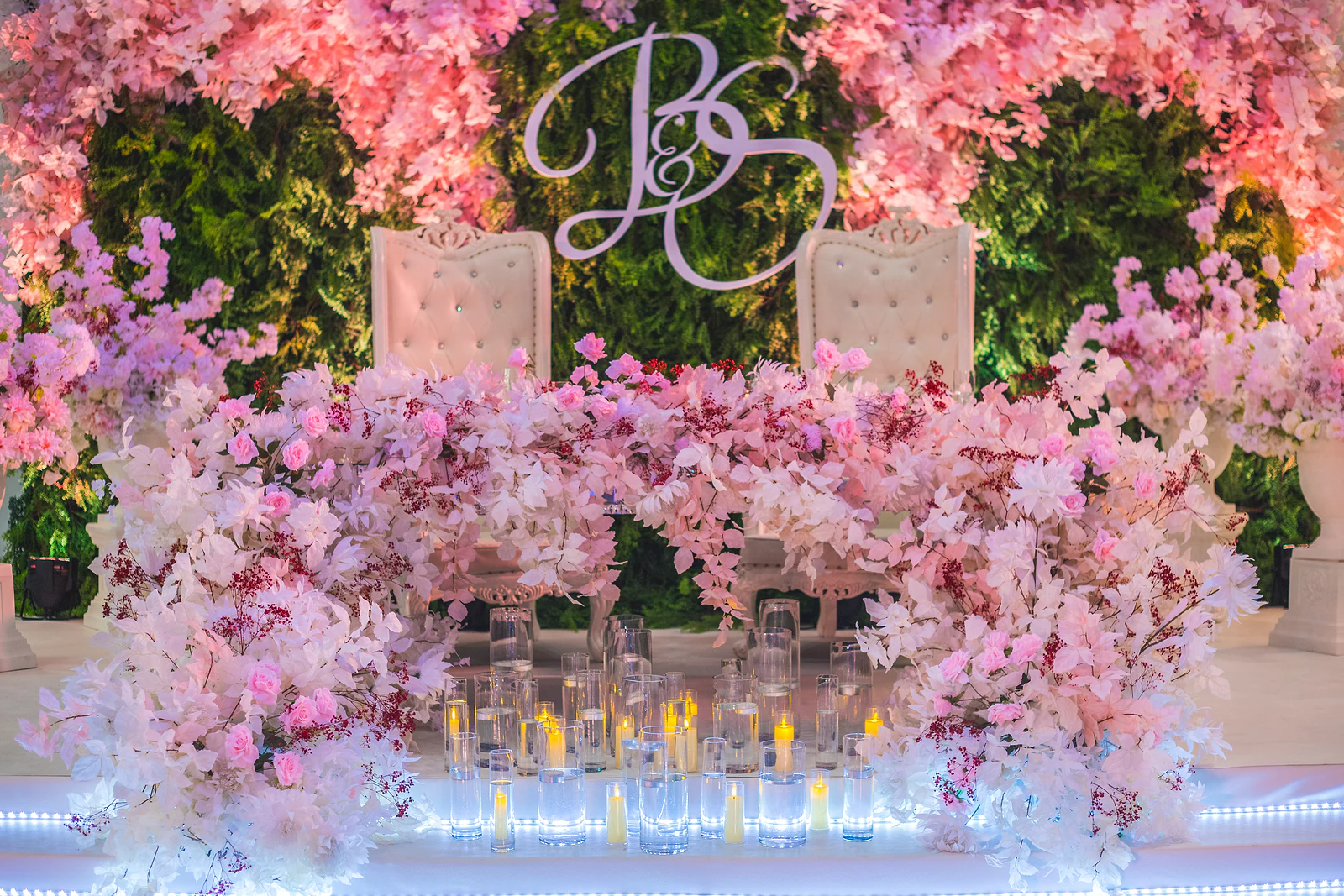 Mesa dos noivos com duas cadeiras estofadas brancas diante de um painel de folhagem com o monograma B&S, rodeada de flores cor-de-rosa e velas em copos de vidro