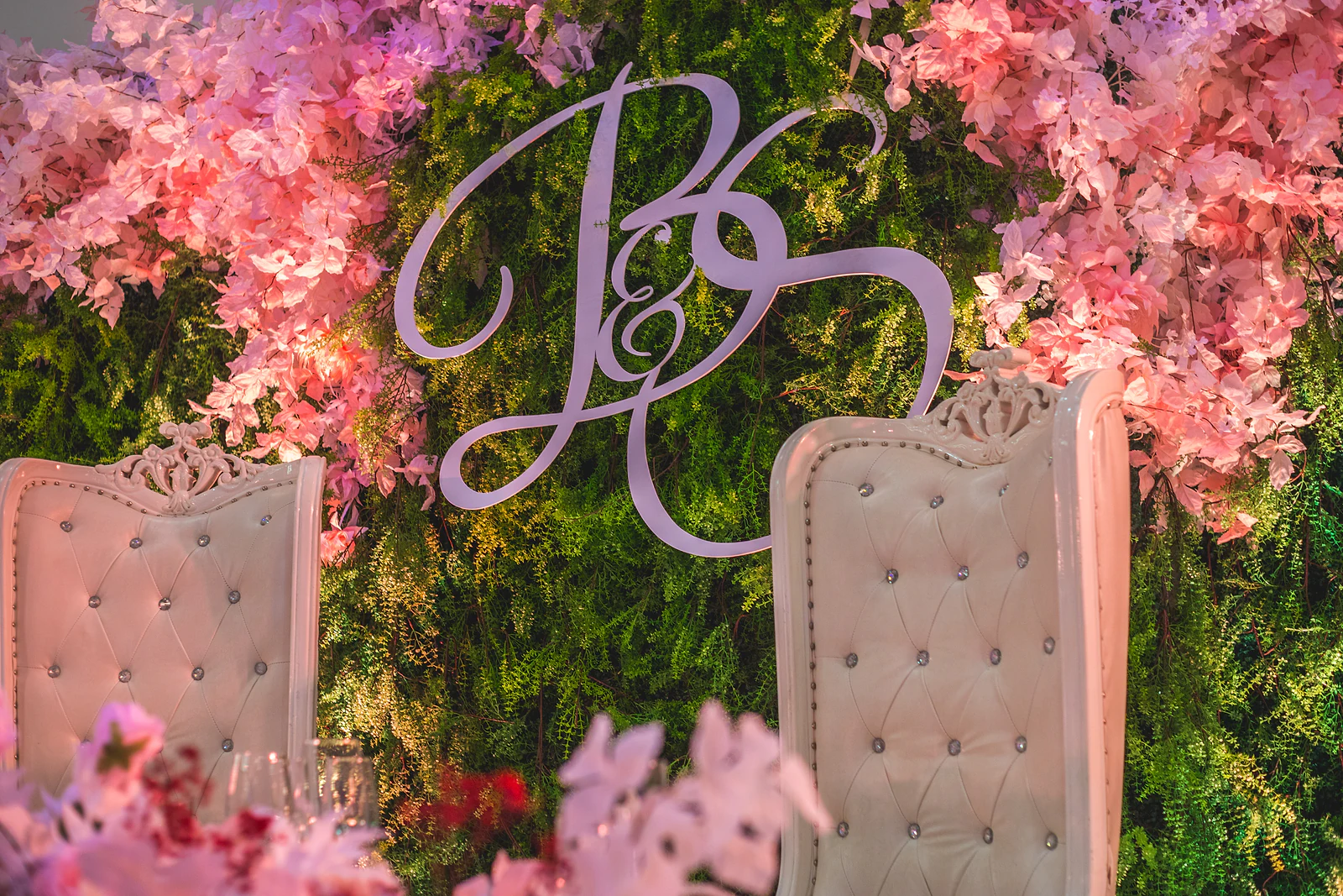 Pormenor das cadeiras altas capitonadas e do monograma prateado sobre a parede de verdura e flores cor-de-rosa