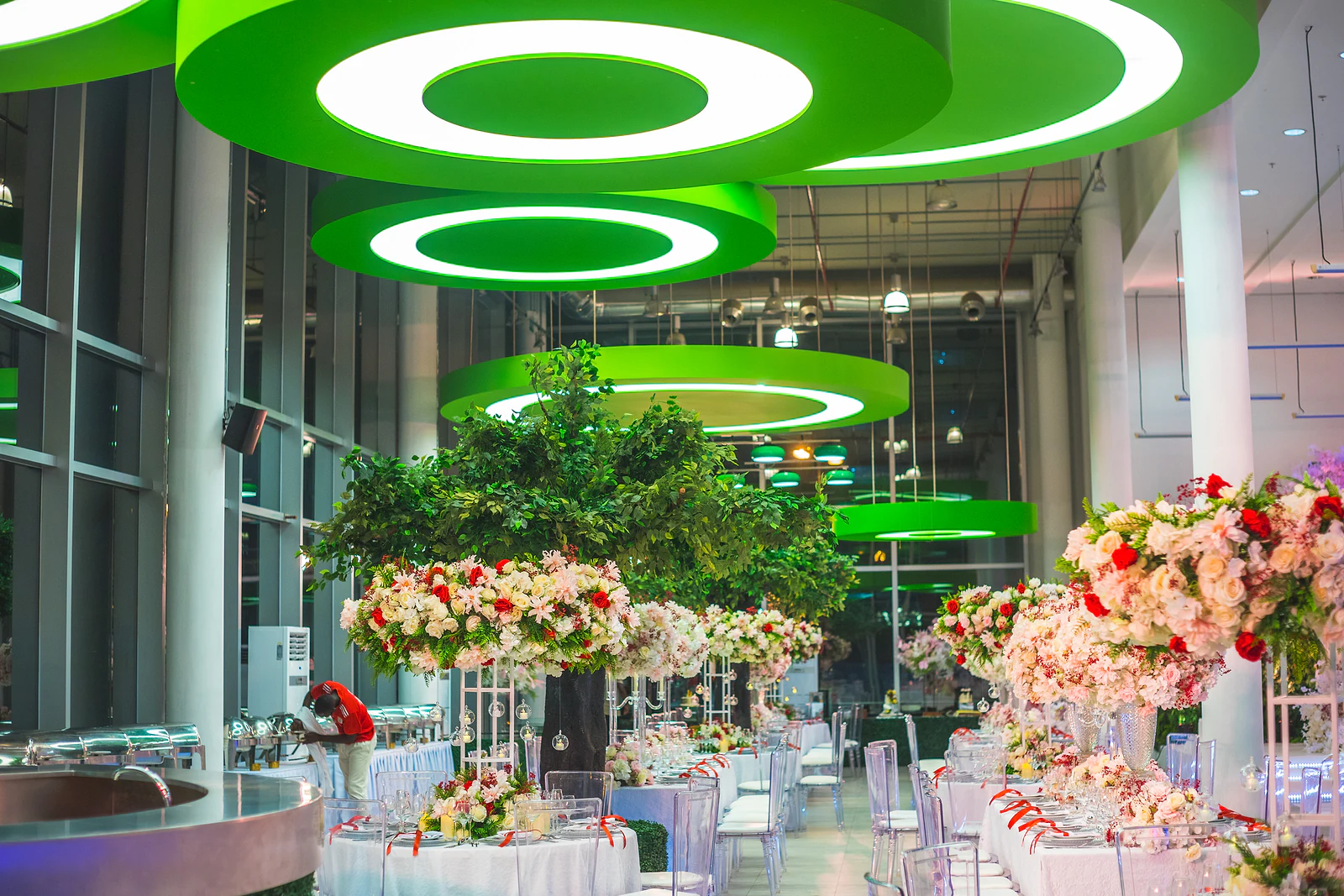 Salão de paredes de vidro com círculos de luz verde no tecto, uma árvore ao centro e mesas com altos centros de flores
