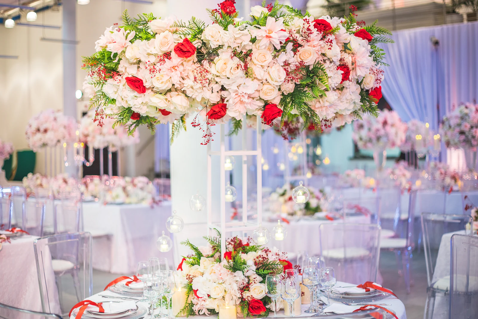 Alto centro de mesa de rosas rosa, brancas e vermelhas sobre um pedestal branco, com mesas postas e cortinados ao fundo
