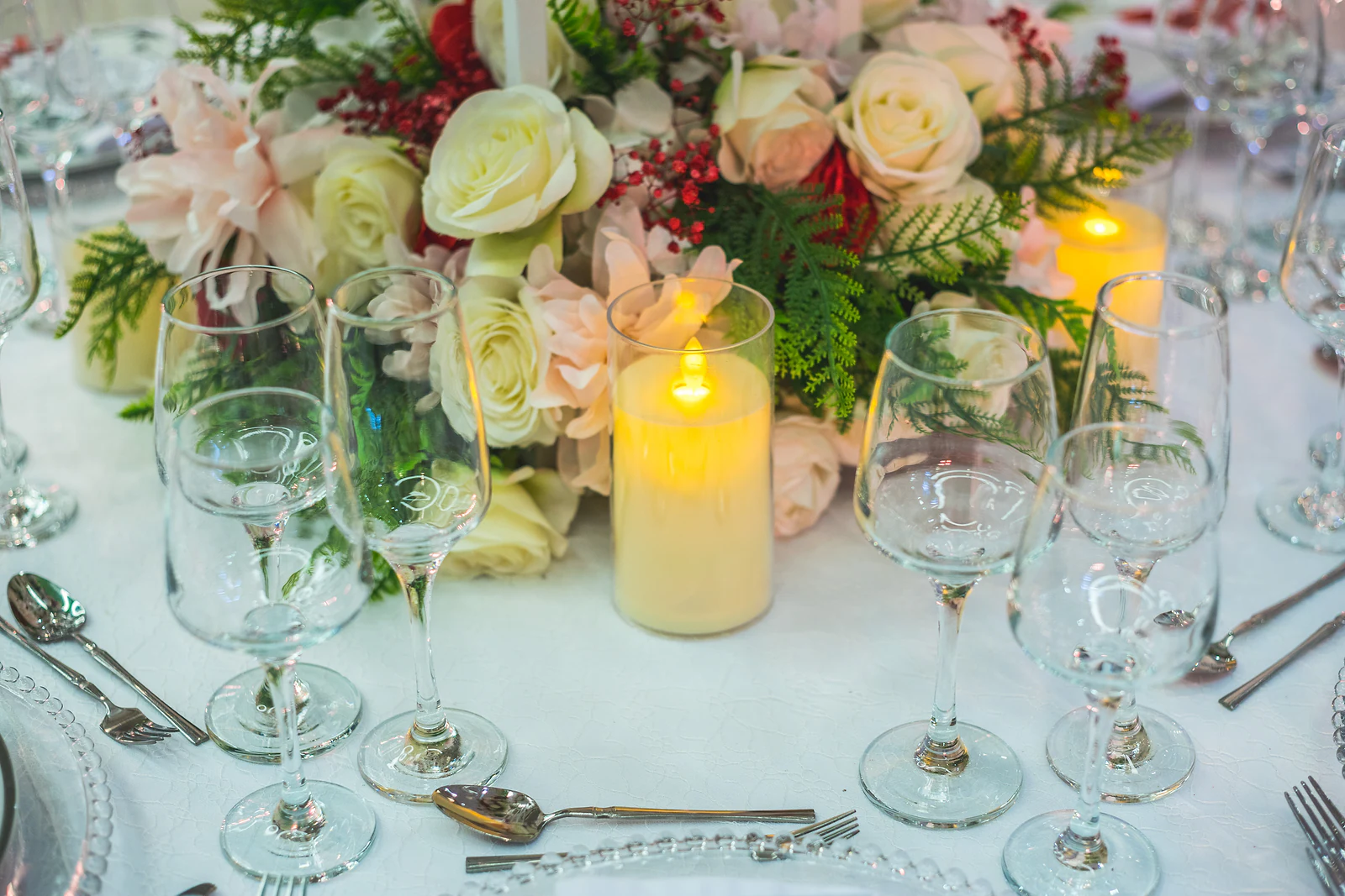 Centro de mesa de rosas cor de marfim, fetos e bagas vermelhas com uma vela acesa e copos de cristal