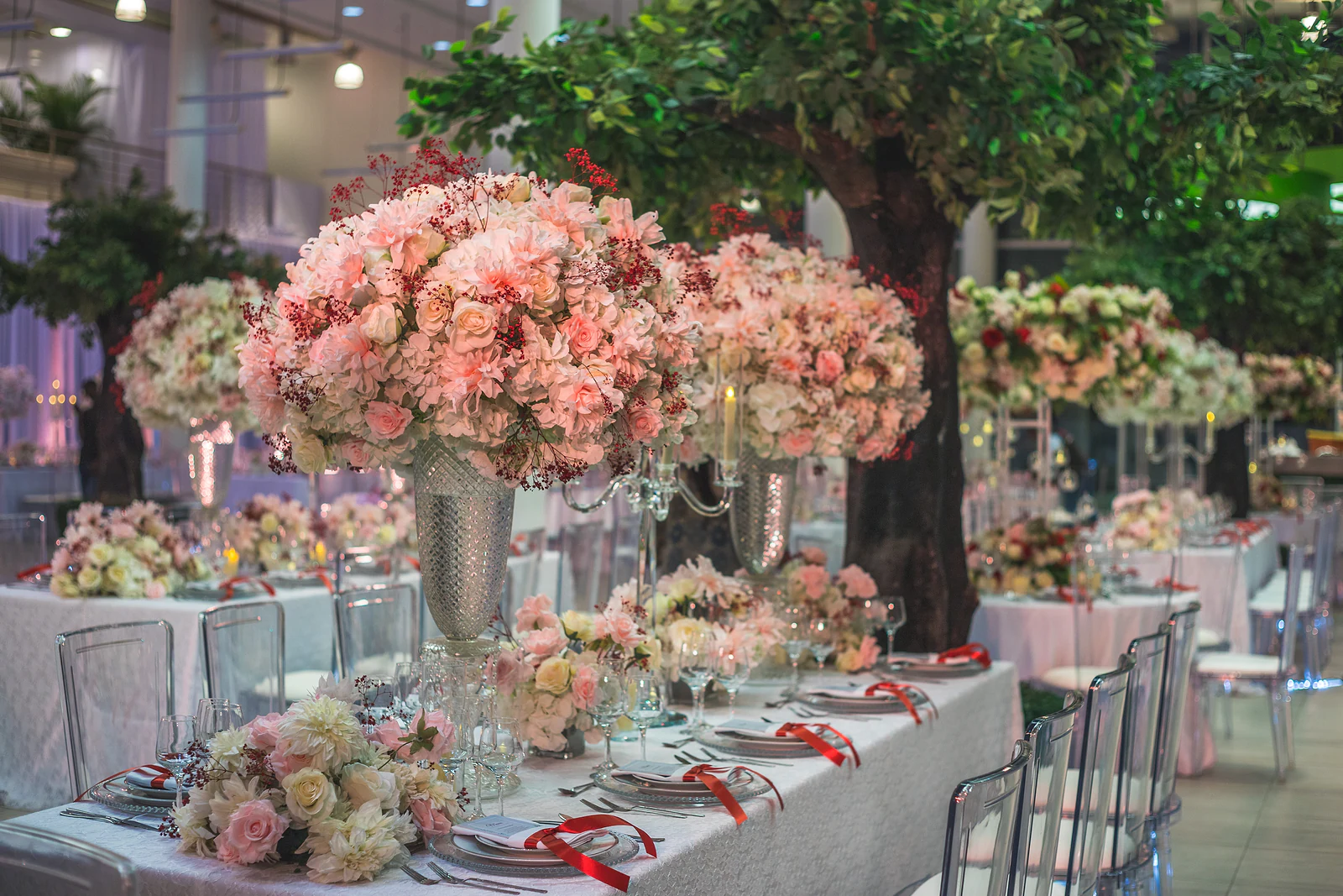 Mesa comprida de toalha branca com cadeiras transparentes e centros de hortênsias rosa, ao lado de uma árvore