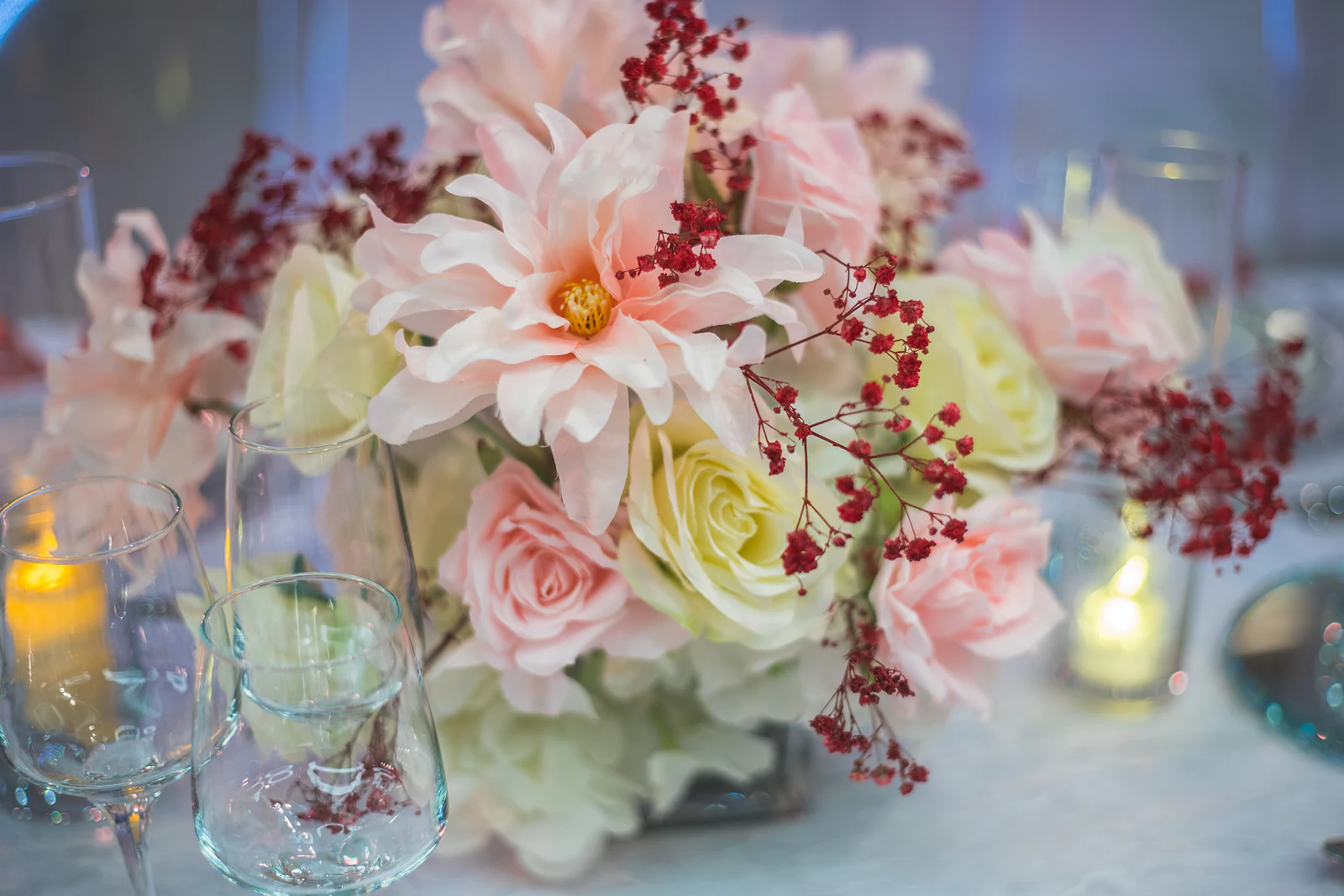 Arranjo de dálias rosa, rosas cor de marfim e bagas vermelhas junto a velas e copos de cristal
