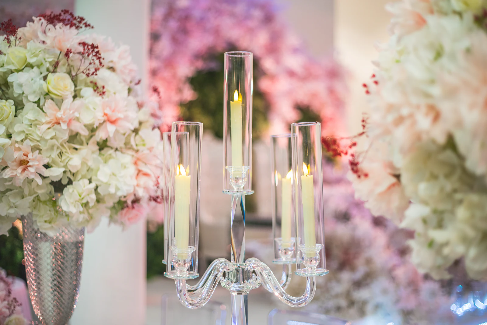 Candelabro de vidro com velas compridas diante de hortênsias brancas e rosa num vaso prateado