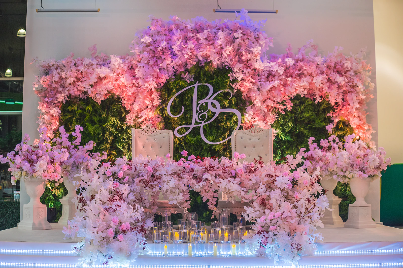 Palco dos noivos com duas cadeiras altas brancas diante de uma parede verde sob arcos de flores cor-de-rosa e o monograma ao centro