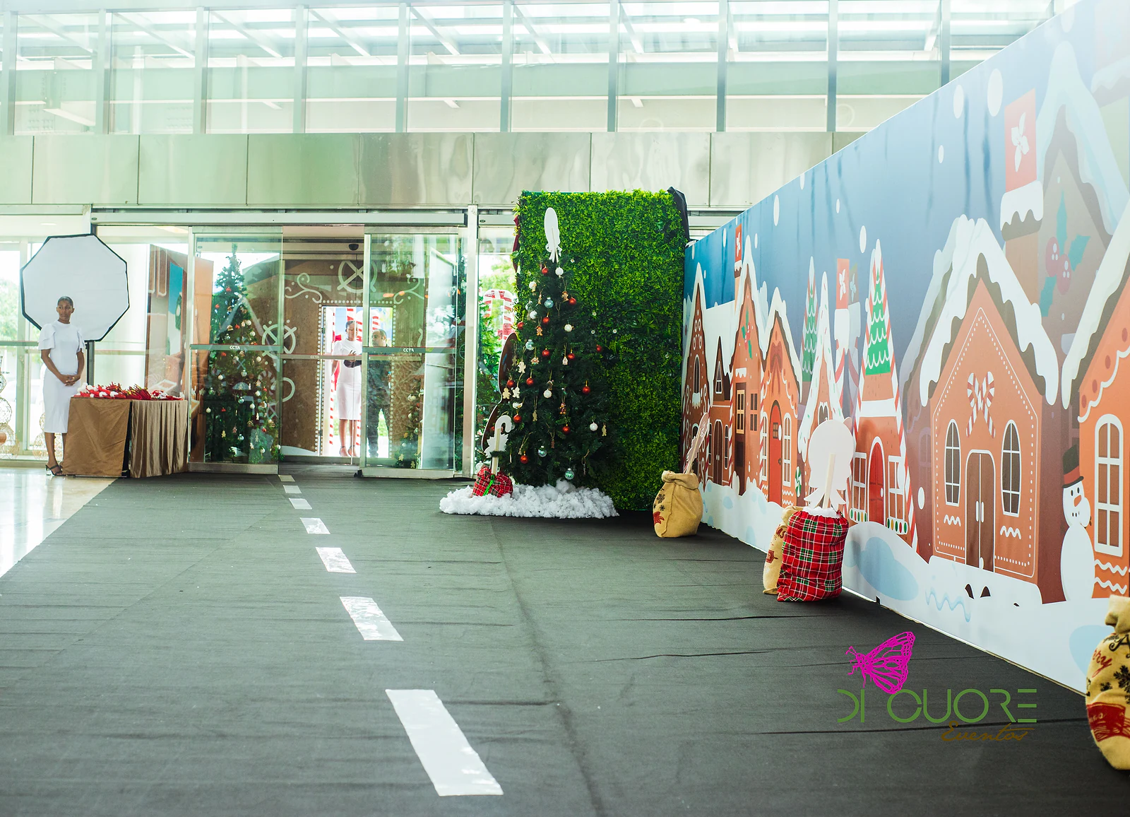 Passadeira preta com marcas de estrada junto a um mural de casinhas de gengibre, com uma árvore de Natal e uma parede de folhagem à entrada