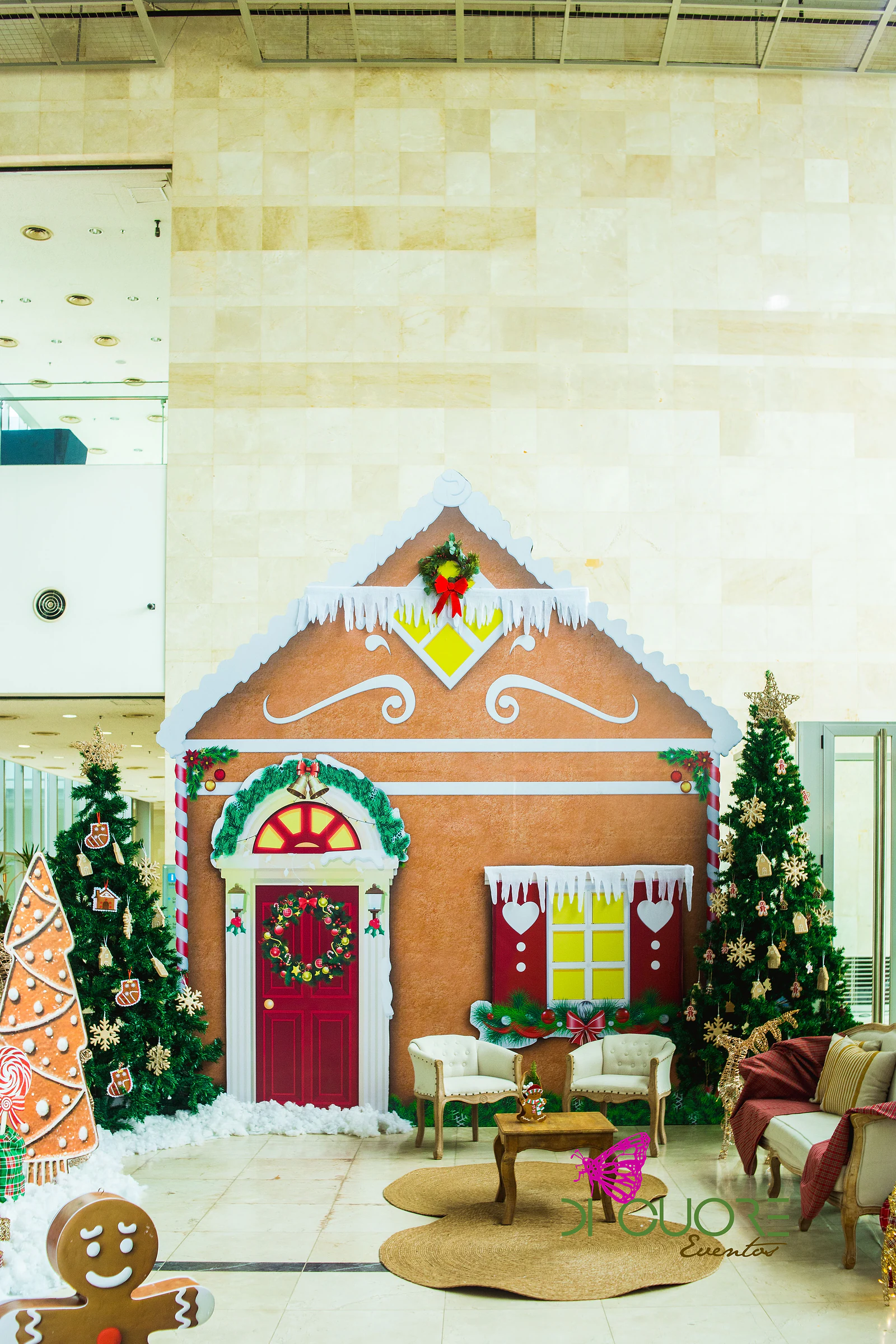 Casa de gengibre com porta vermelha e coroa de Natal, entre árvores de Natal, duas poltronas brancas e uma mesa de madeira sobre um tapete de corda