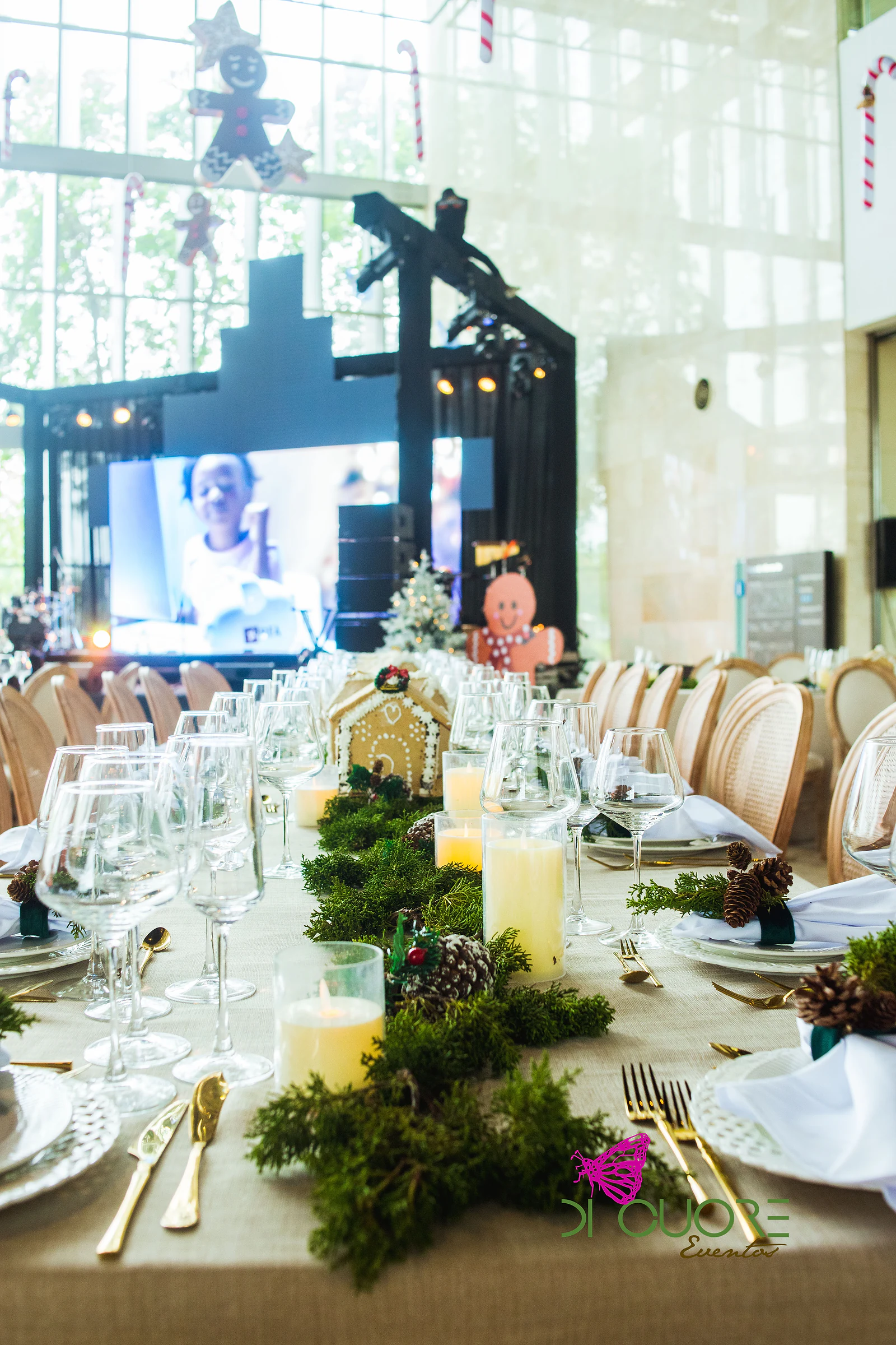 Mesa posta com casinha de gengibre, musgo, pinhas e velas entre copos de cristal, com o palco e o ecrã ao fundo