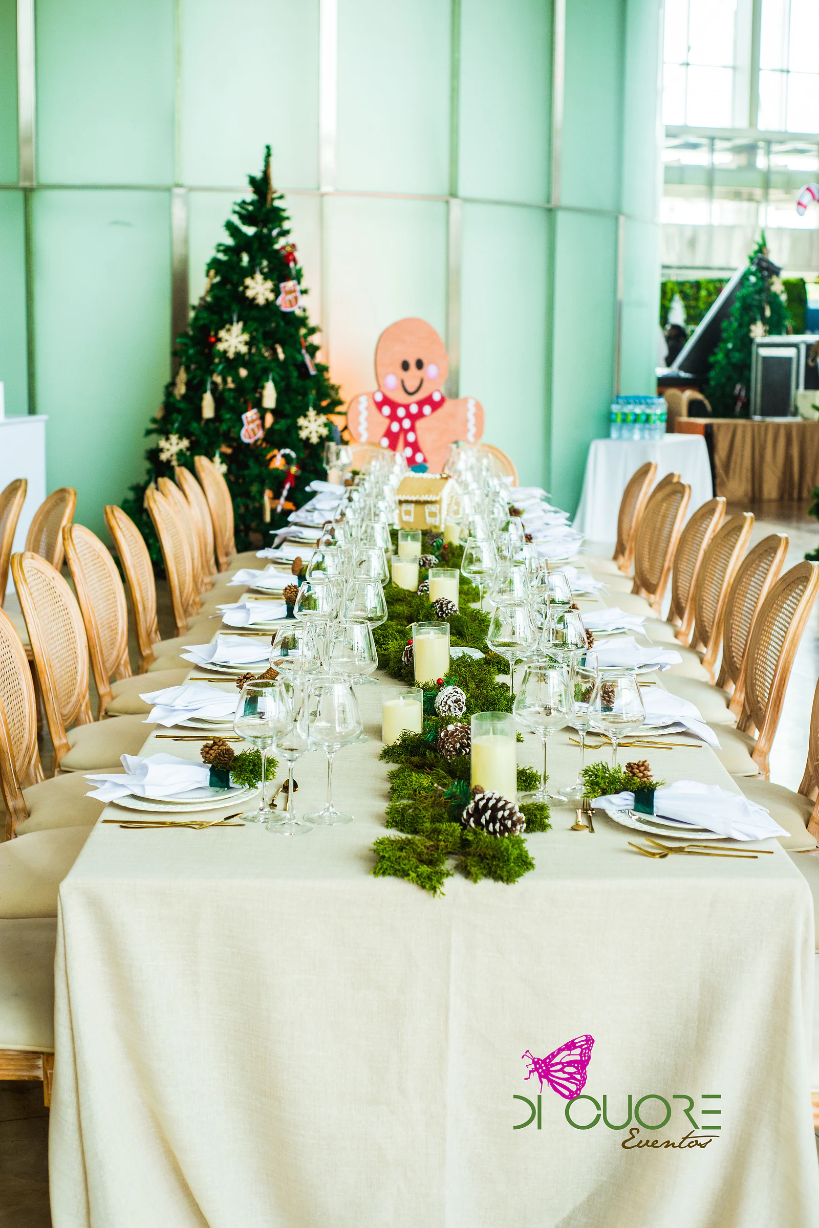 Mesa comprida de linho cru com carreiro de musgo, pinhas e velas, cadeiras de palhinha e, ao fundo, uma árvore de Natal e um boneco de gengibre