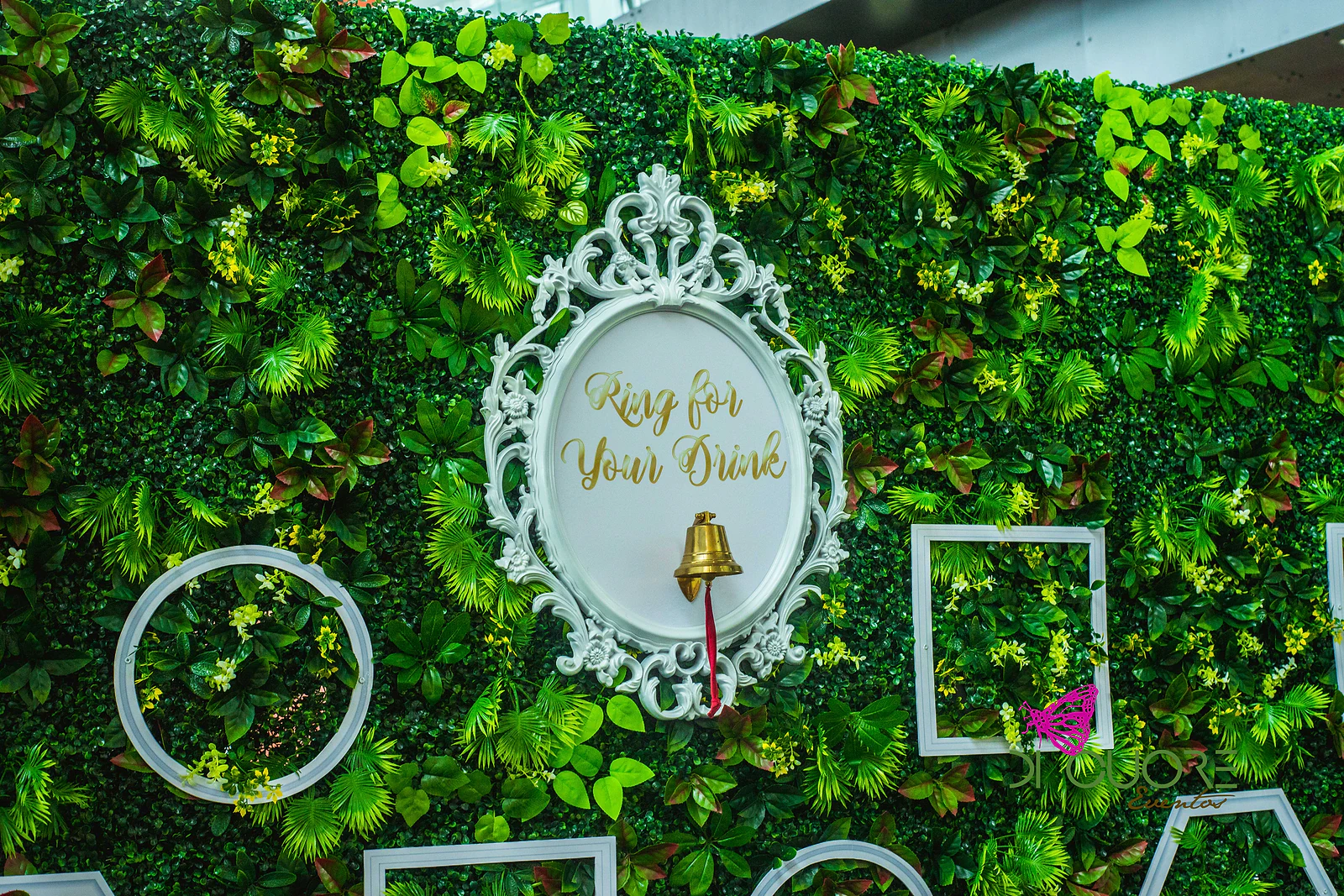 Parede de folhagem com moldura branca ornamentada, sineta dourada e a frase “Ring for Your Drink”