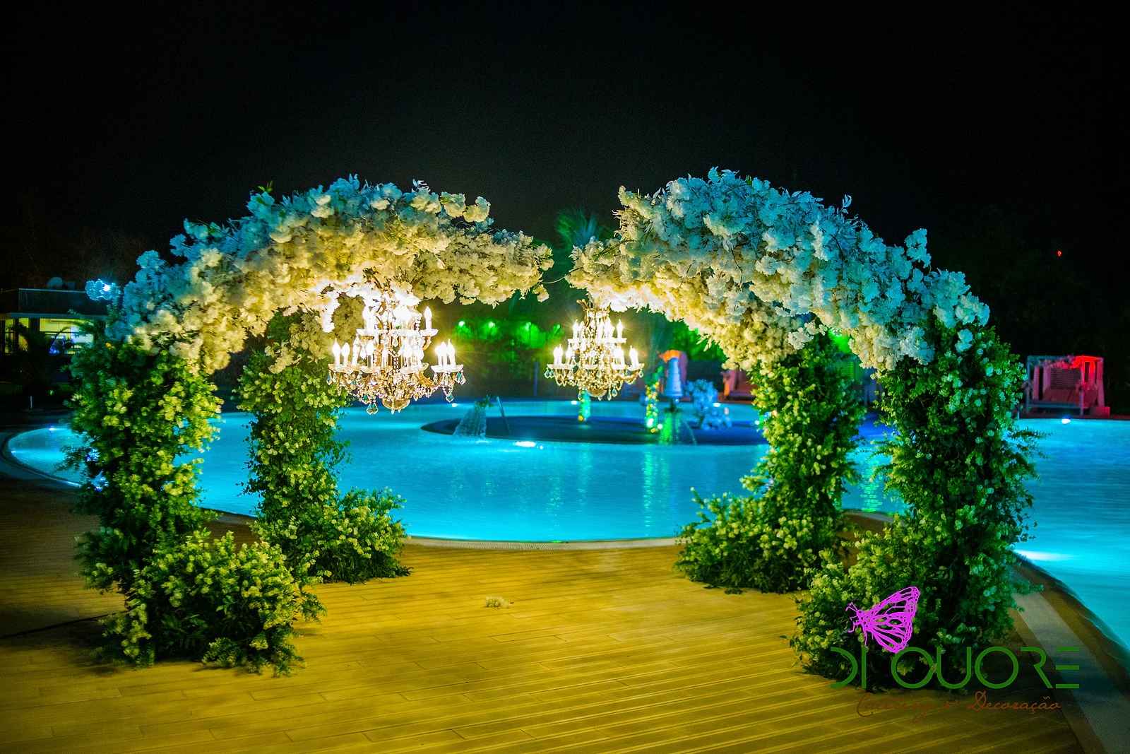 Dois arcos de flores brancas e folhagem iluminados à noite, com lustres de cristal, sobre a piscina acesa em turquesa