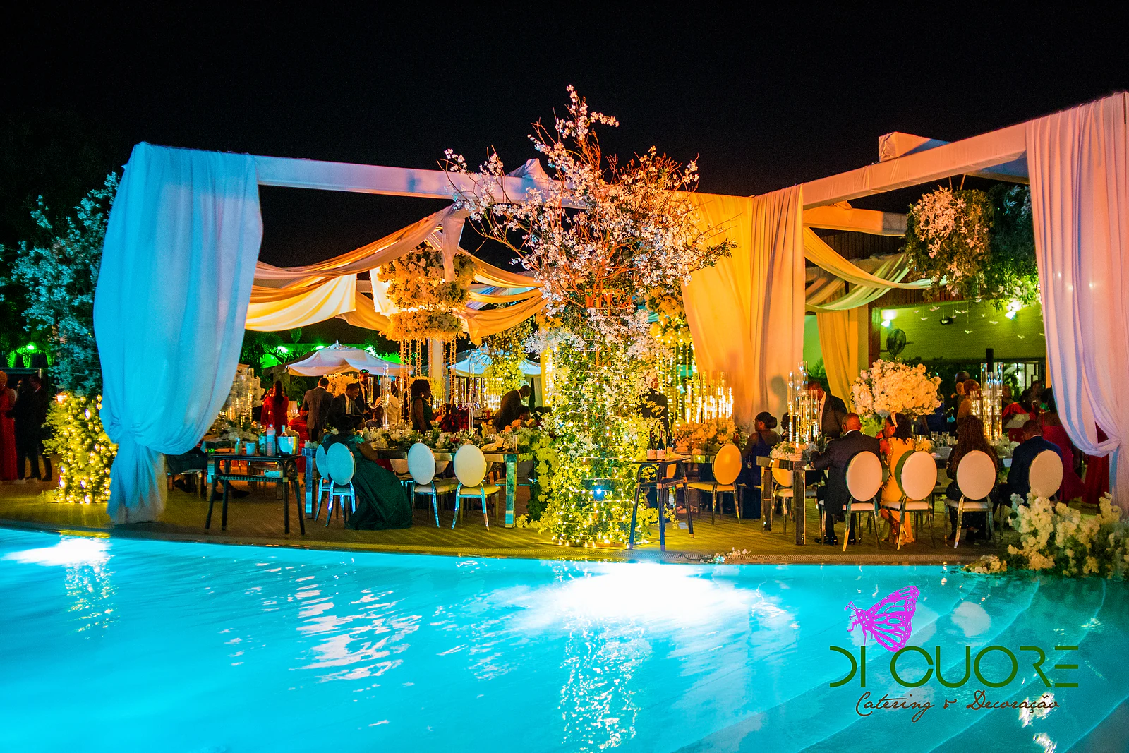 Pérgolas com cortinados rosa e brancos sobre as mesas, iluminadas à noite, com um arbusto de flores no centro e a piscina em primeiro plano