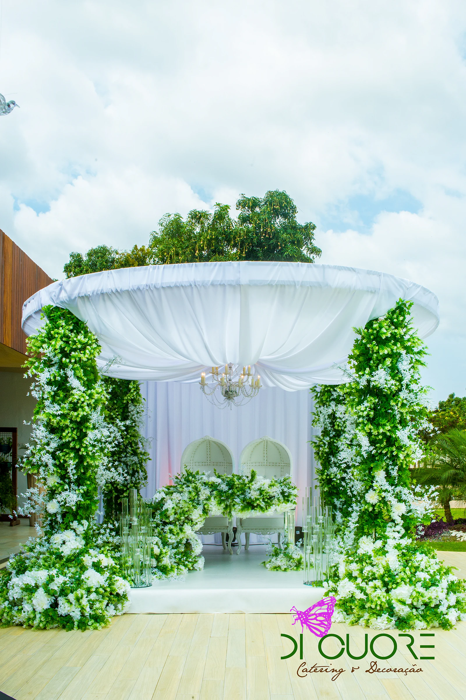 Pavilhão com cobertura de tecido branco entre duas colunas de folhagem e flores brancas, com dois cadeirões brancos ao fundo, sobre um deque de madeira clara