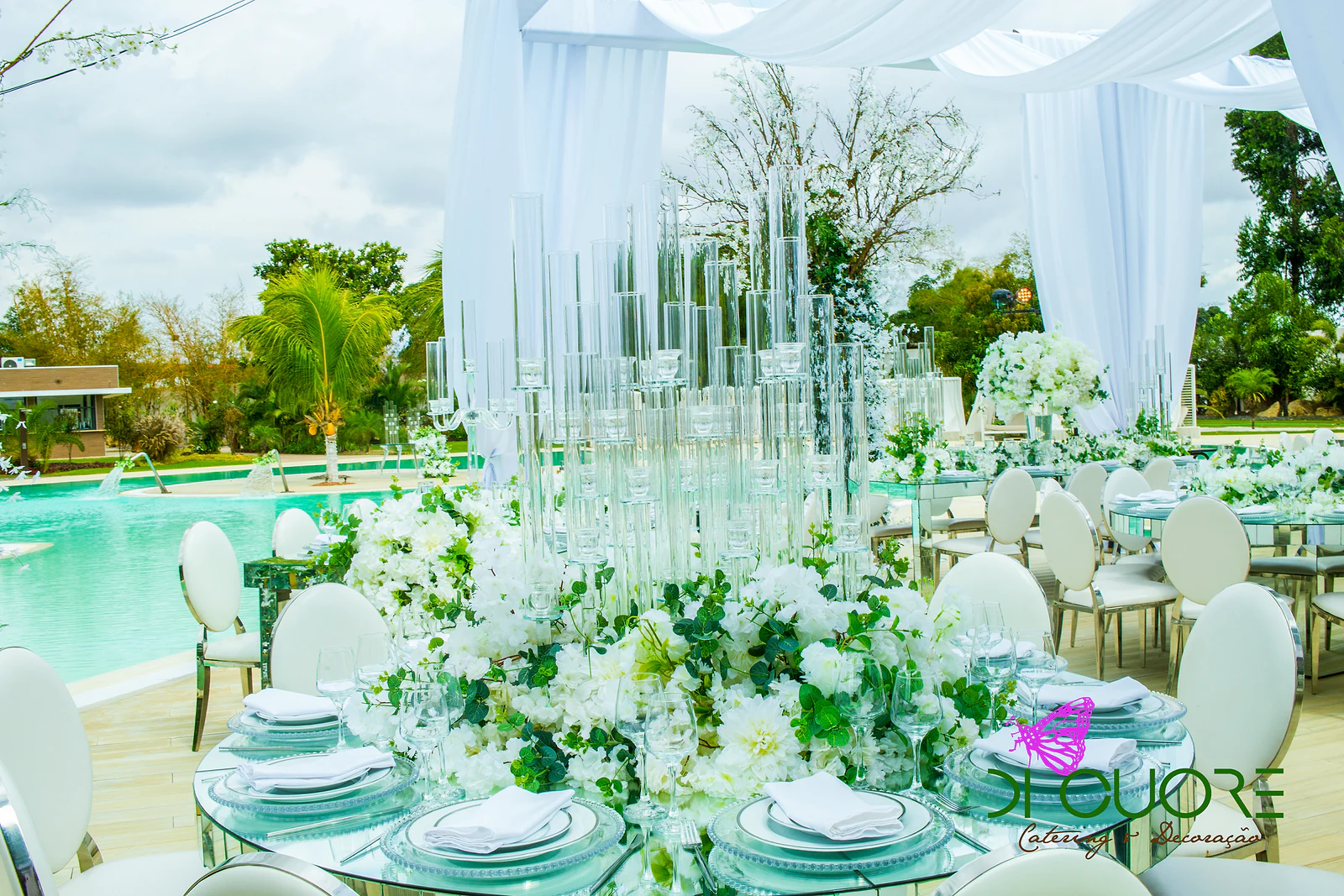 Mesa redonda de tampo espelhado com pratos azul-claros, flores brancas e folhagem, e um centro de mesa de tubos de cristal, junto à piscina