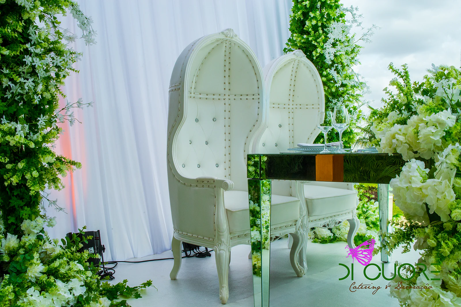 Dois cadeirões brancos de espaldar alto e botões de cristal, junto a uma mesa de espelhos, entre cortinado branco e colunas de flores