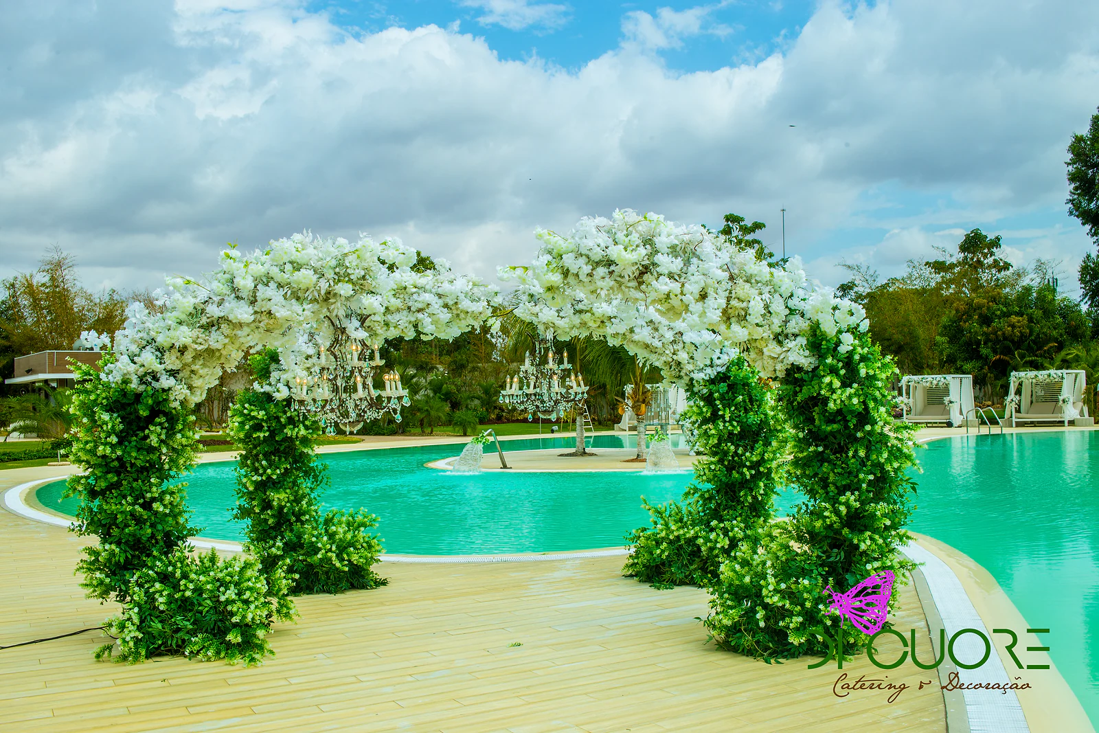 Dois arcos de flores brancas apoiados em colunas de folhagem sobre o deque, com lustres suspensos e a piscina turquesa ao fundo
