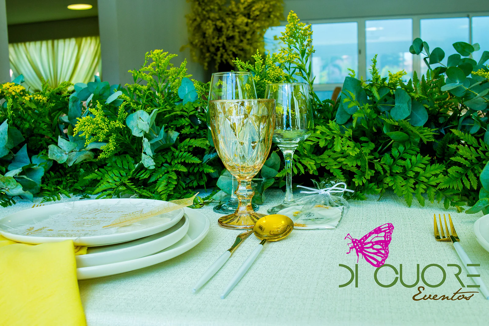 Caminho de mesa de folhagem, eucalipto e flores amarelas, com um copo dourado, uma colher dourada e uma lembrança em saquinho transparente