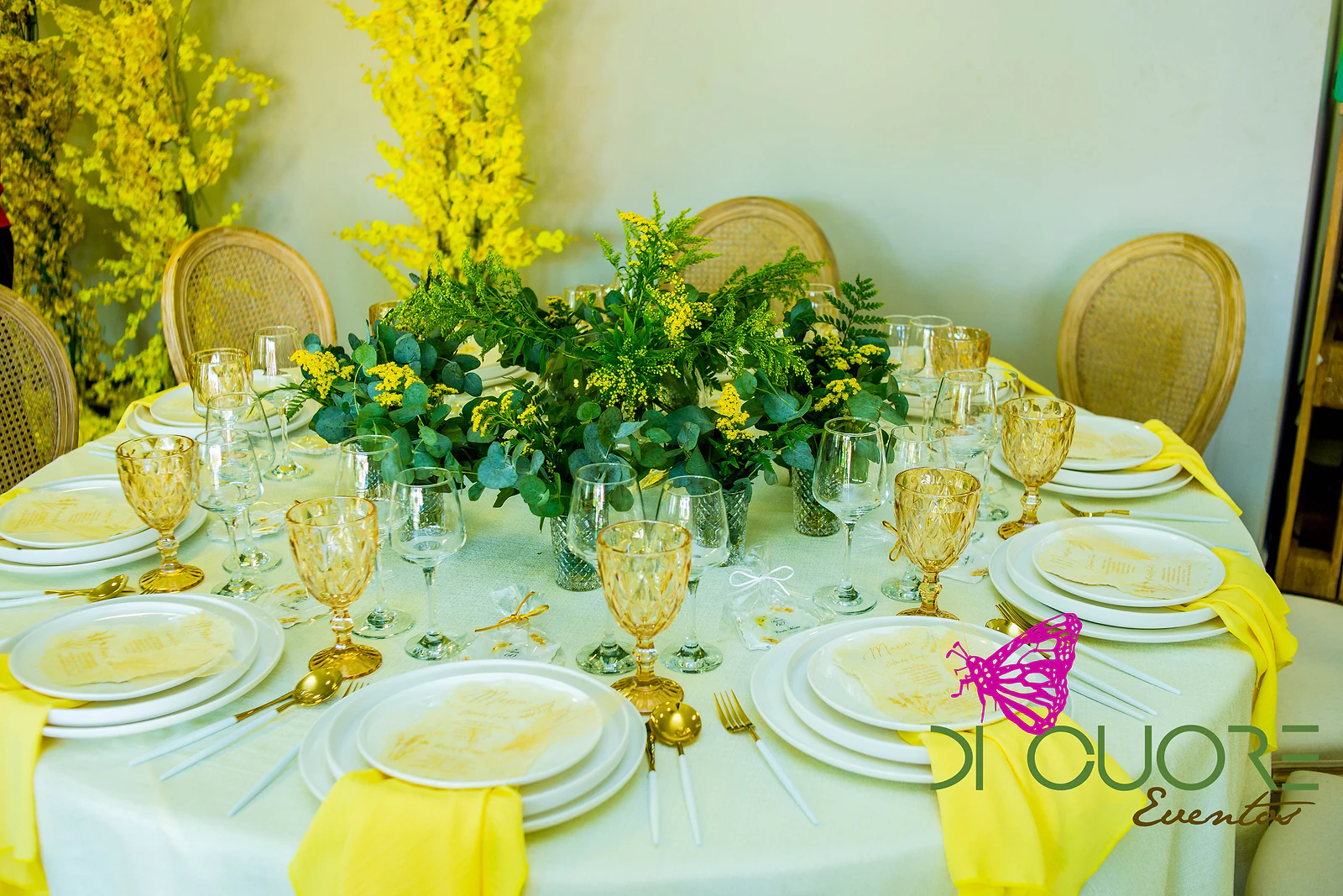 Mesa redonda de toalha clara com guardanapos amarelos, copos cor de âmbar, pratos brancos e um centro de folhagem e flores amarelas, rodeada de cadeiras de palhinha