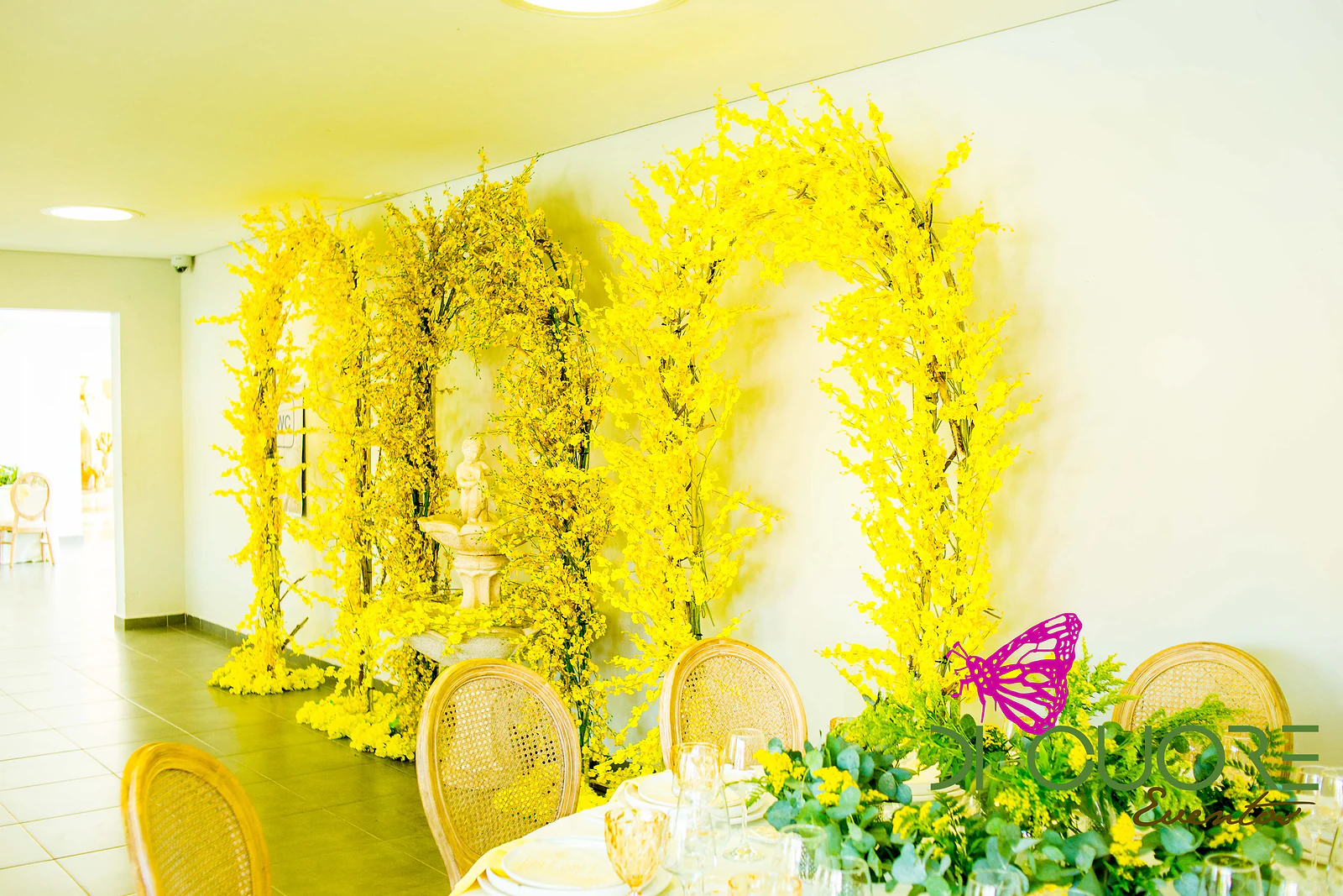 Arcos de flores amarelas à volta de uma fonte de pedra, ao fundo, com uma mesa posta e cadeiras de palhinha em primeiro plano