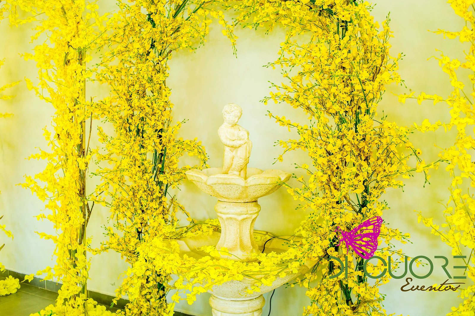 Fonte de pedra clara com uma figura de menino, rodeada de ramos de flores amarelas, junto a uma parede amarelada