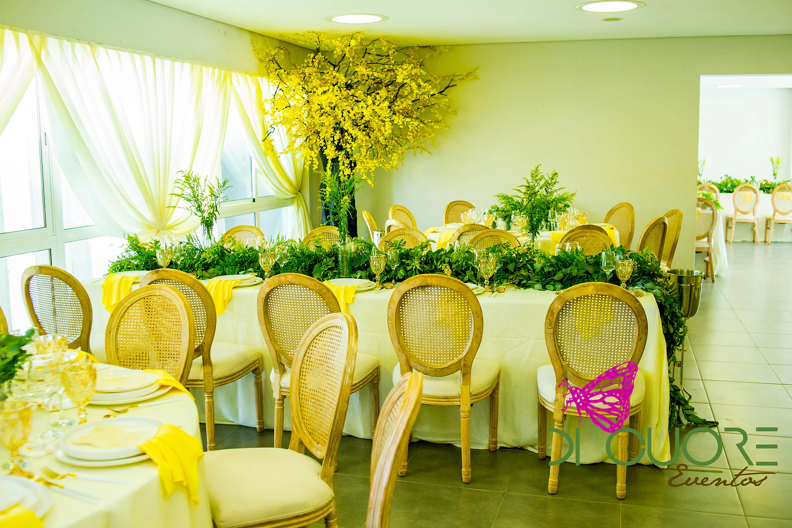 Mesas postas com caminho de folhagem e cadeiras de madeira com palhinha, sob uma grande árvore de flores amarelas e cortinados claros