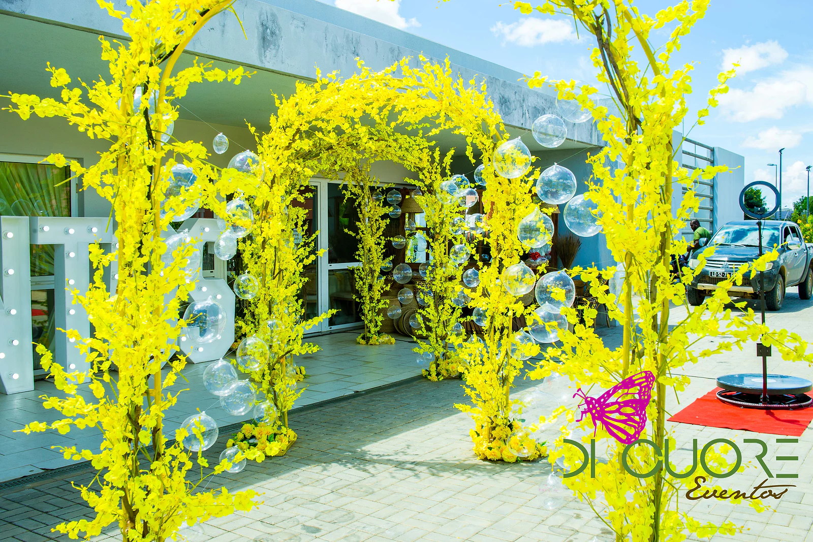 Fila de arcos de flores amarelas com esferas de acrílico transparentes à porta do salão, com as grandes letras brancas ao lado