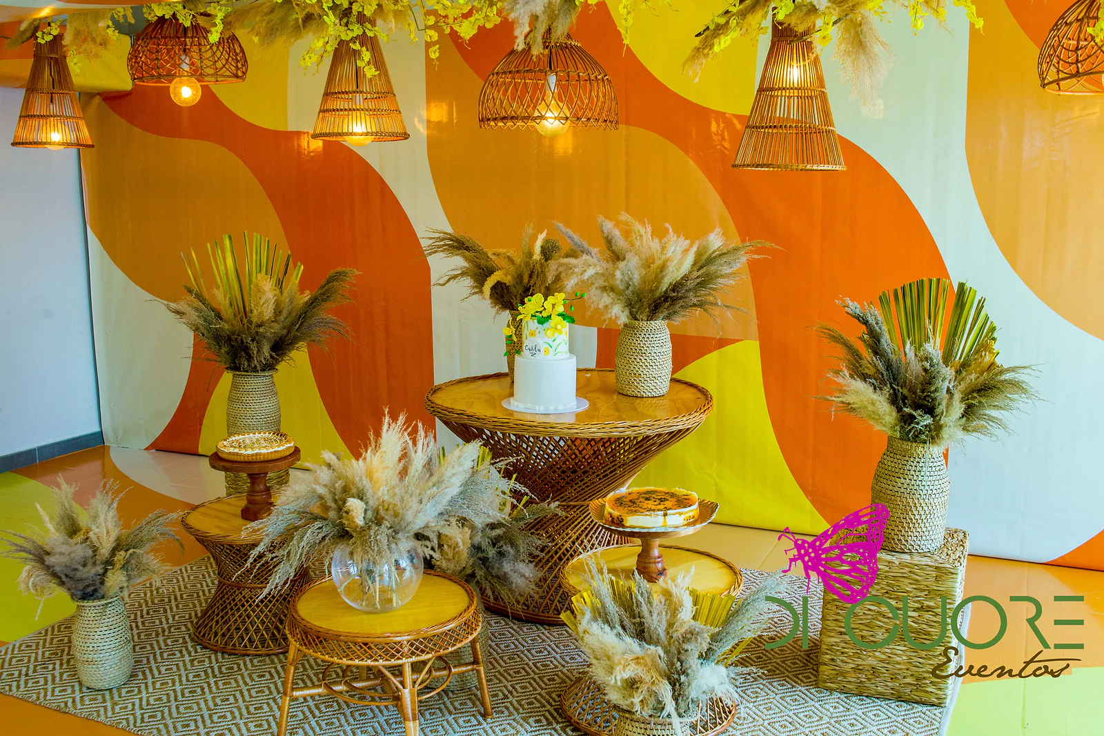 Mesa do bolo diante de um painel de ondas laranja e amarelas, com bolo branco de flores amarelas, doces em bancos de vime, vasos de palha com ervas-das-pampas e candeeiros de vime