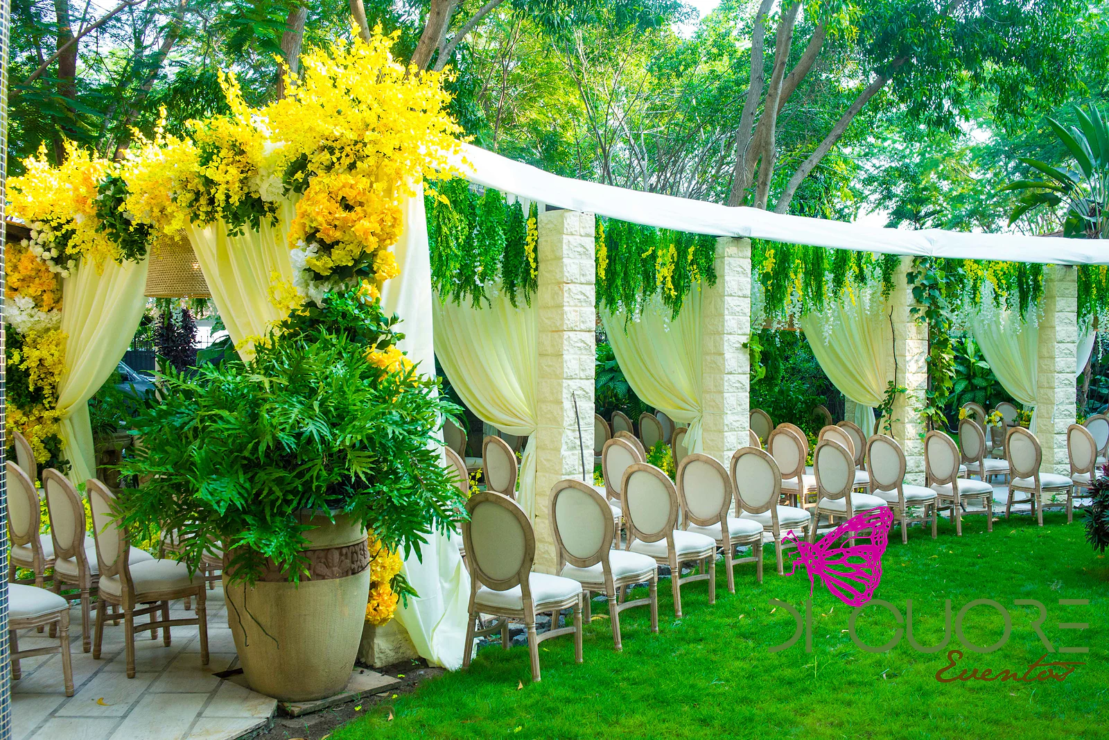 Row of pale wooden chairs on the lawn, beneath a white fabric canopy between stone pillars with trailing foliage, beside the portal of yellow orchids
