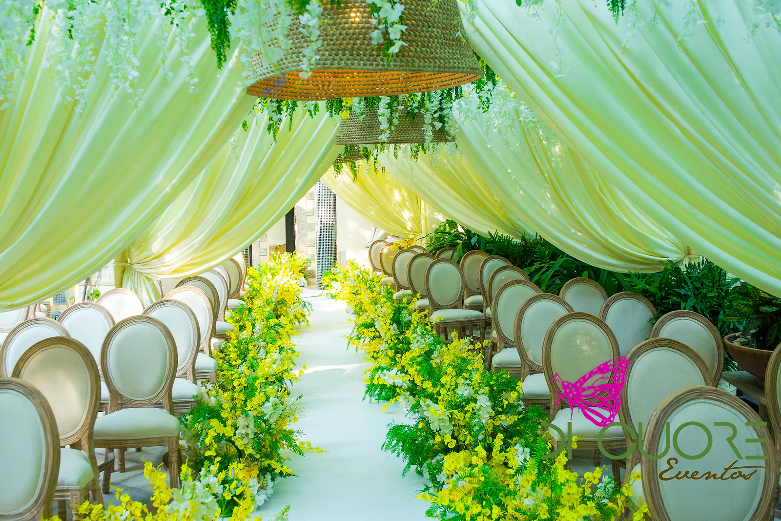 White aisle between rows of pale wooden chairs, edged with yellow orchids and ferns, beneath butter-coloured drapes and straw lamps