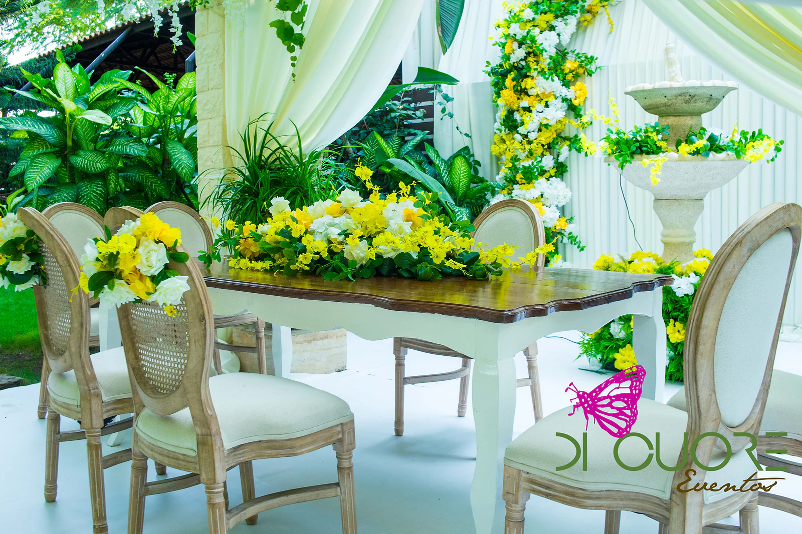 Table with a dark wooden top and white legs and a low arrangement of yellow and white flowers, pale wooden chairs and a stone fountain behind