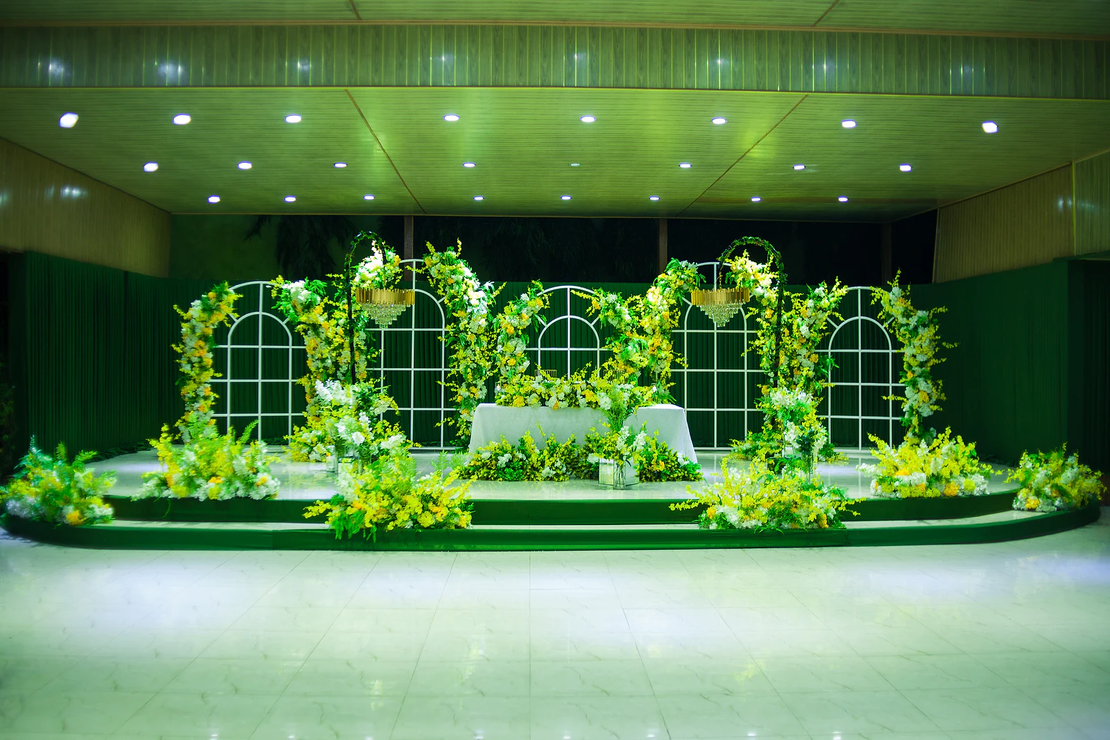 Stage with a white-clothed table before window-shaped panels covered in yellow and white flowers, with chandeliers and green-covered steps