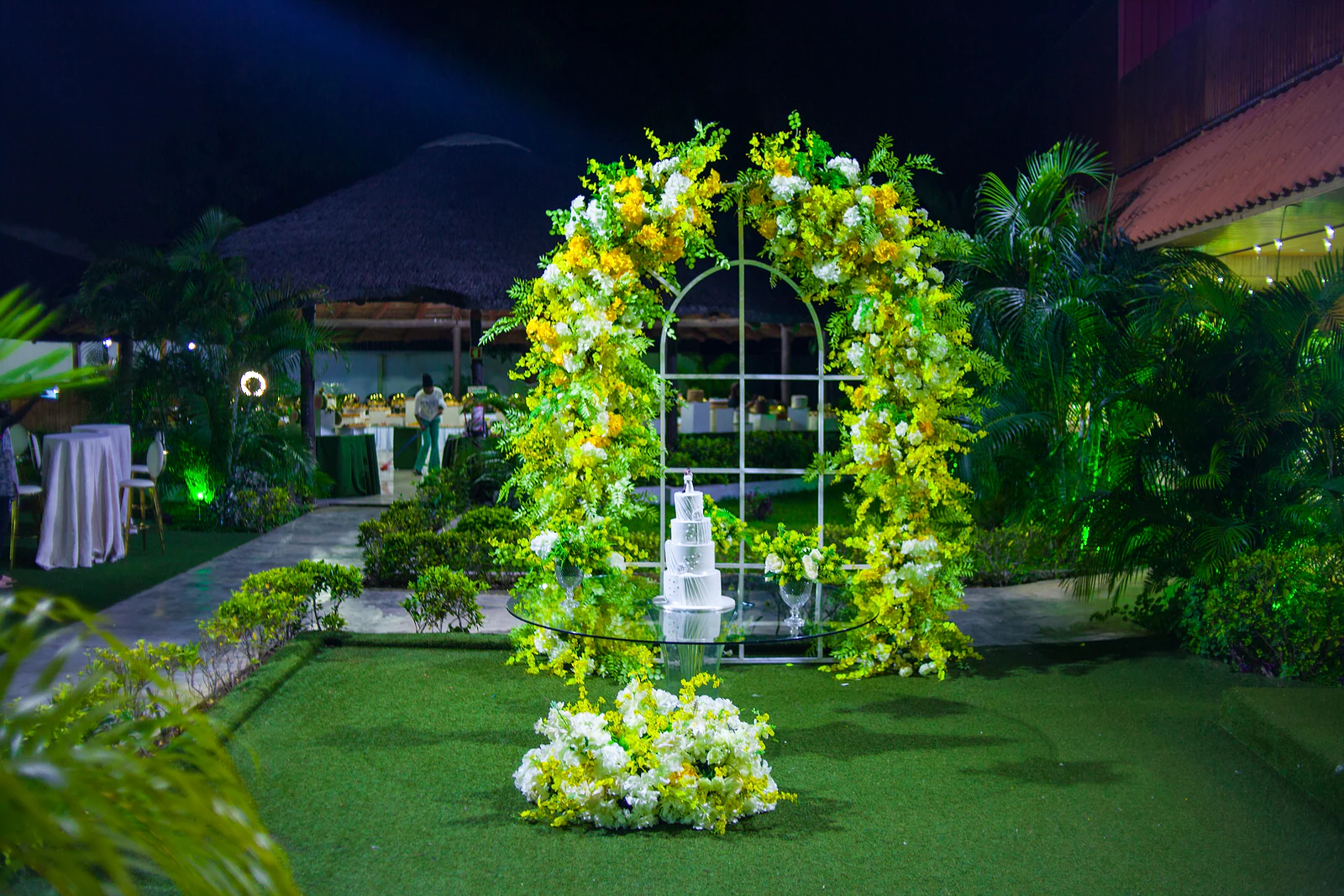 Arch of yellow and white flowers with a window-shaped frame around the cake on a glass table, in the garden at night