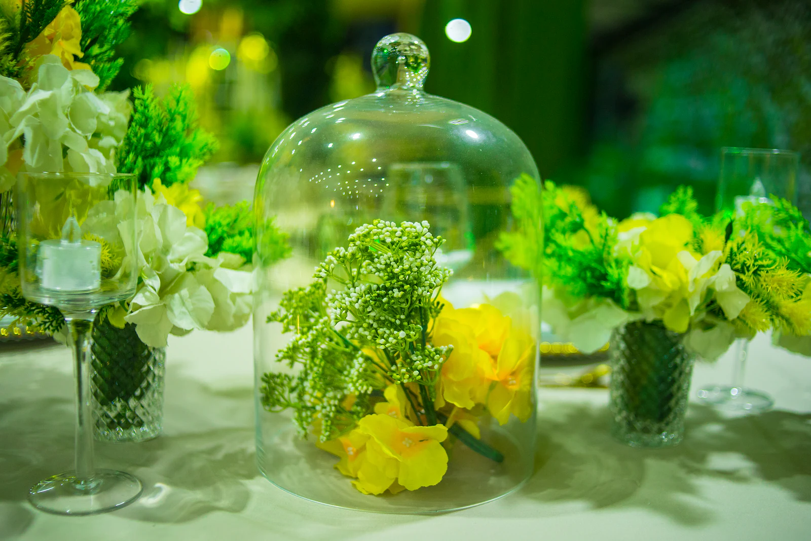 Glass cloche with gypsophila and yellow flowers, between small yellow and white arrangements and a candle in a glass holder