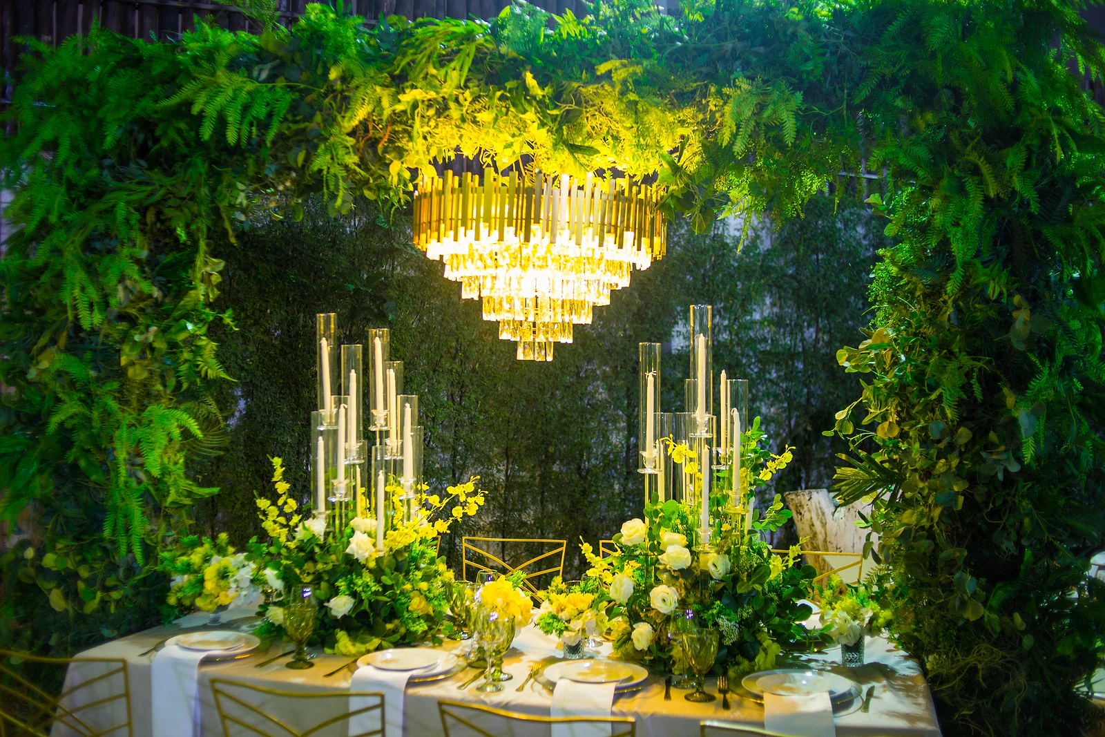 Laid table beneath a foliage portal and a golden chandelier, with candle candelabra and arrangements of yellow and white flowers