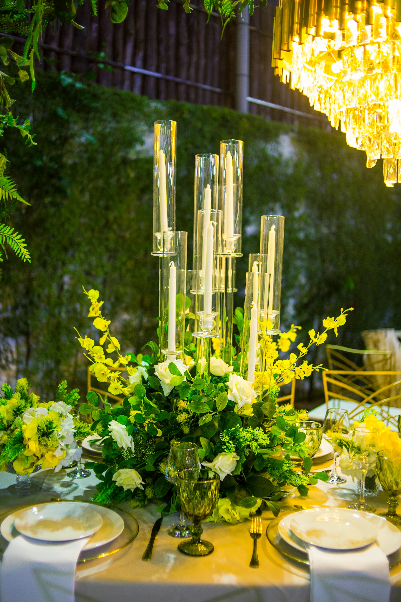 Table centrepiece of candle candelabra, foliage and yellow and white flowers, beneath a golden chandelier
