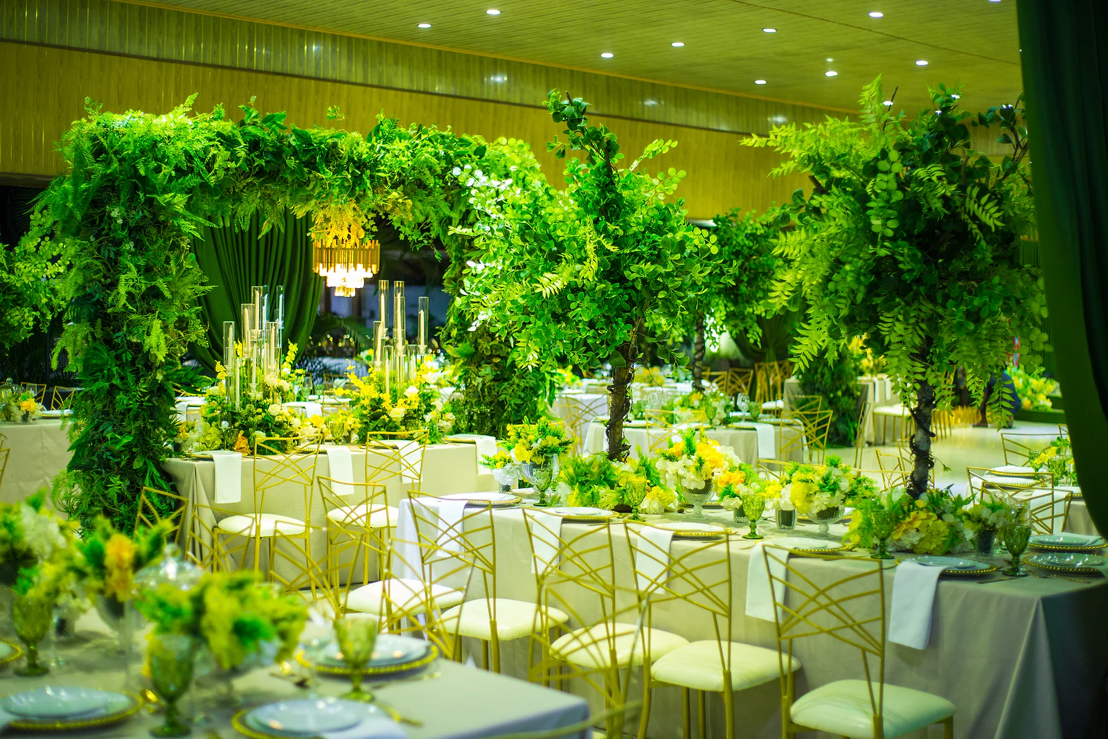Hall with laid tables in grey cloths and gold chairs, leafy trees and a green foliage portal beyond