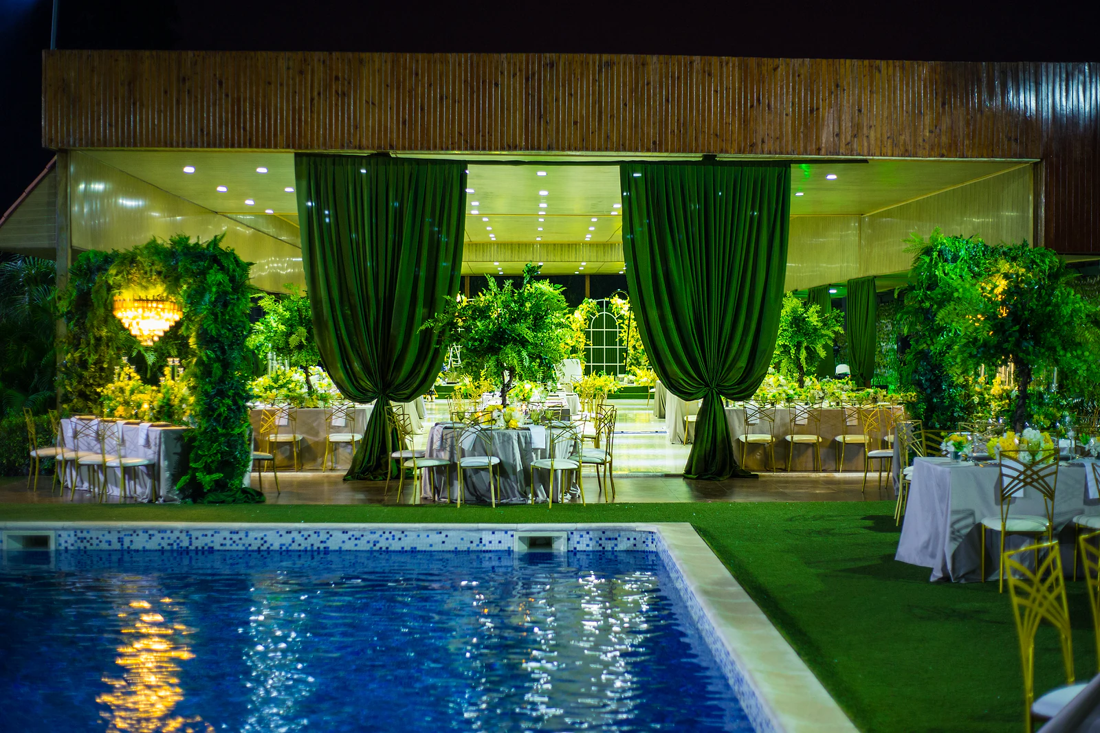 Pool lit at night before the hall, with green velvet drapes, leafy trees and laid tables