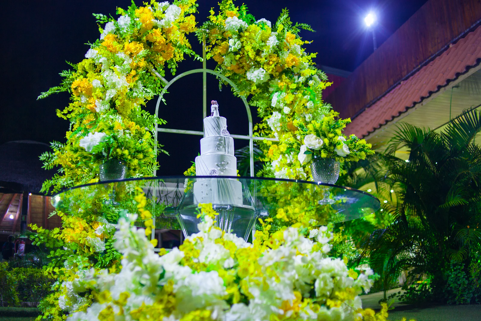 White four-tier cake with the couple’s topper, on a glass table beneath an arch of yellow and white flowers and foliage