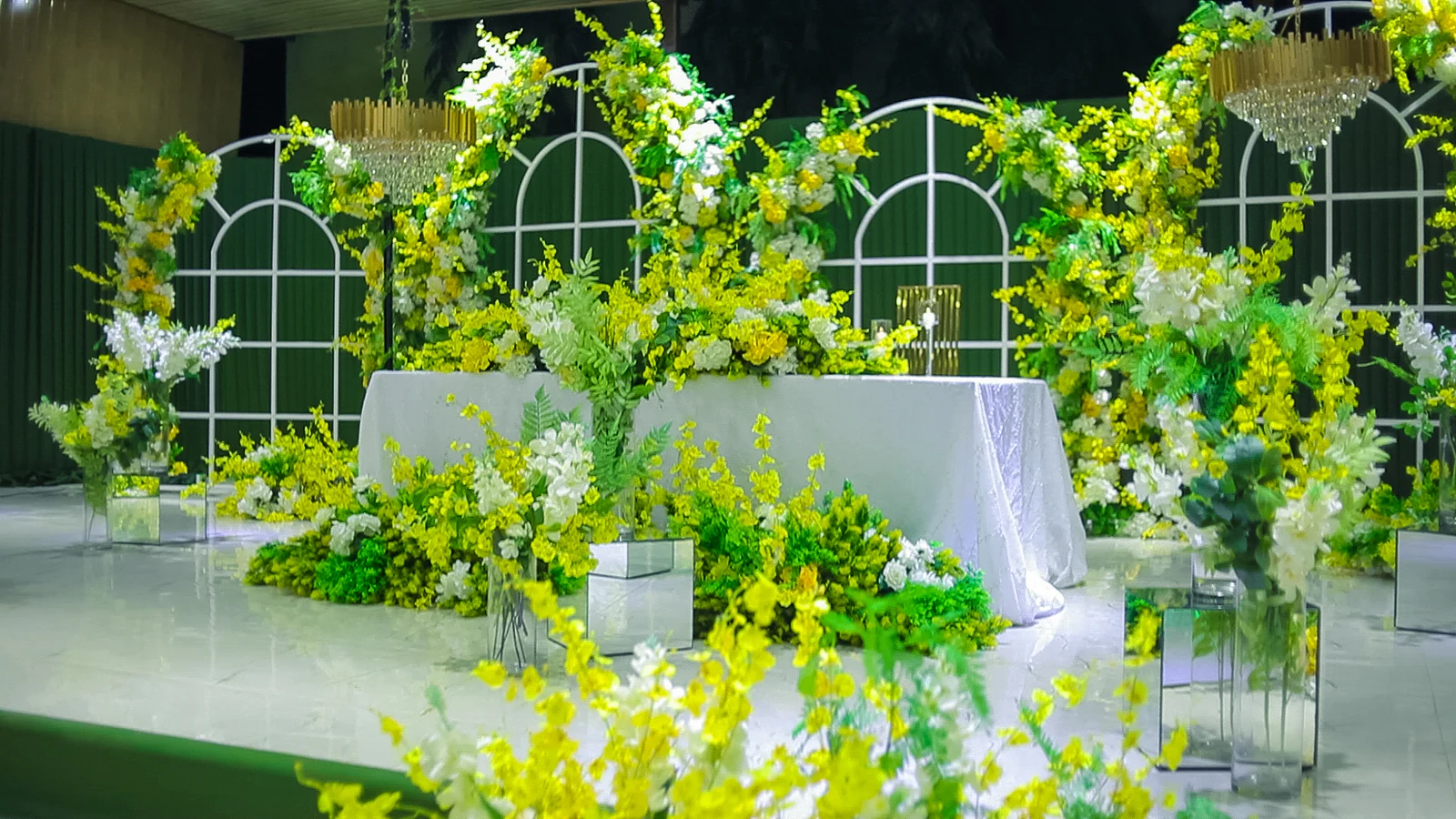 Long table with a white cloth before arched windows on a green backdrop, surrounded by yellow orchids, foliage and white flowers, with golden crystal chandeliers