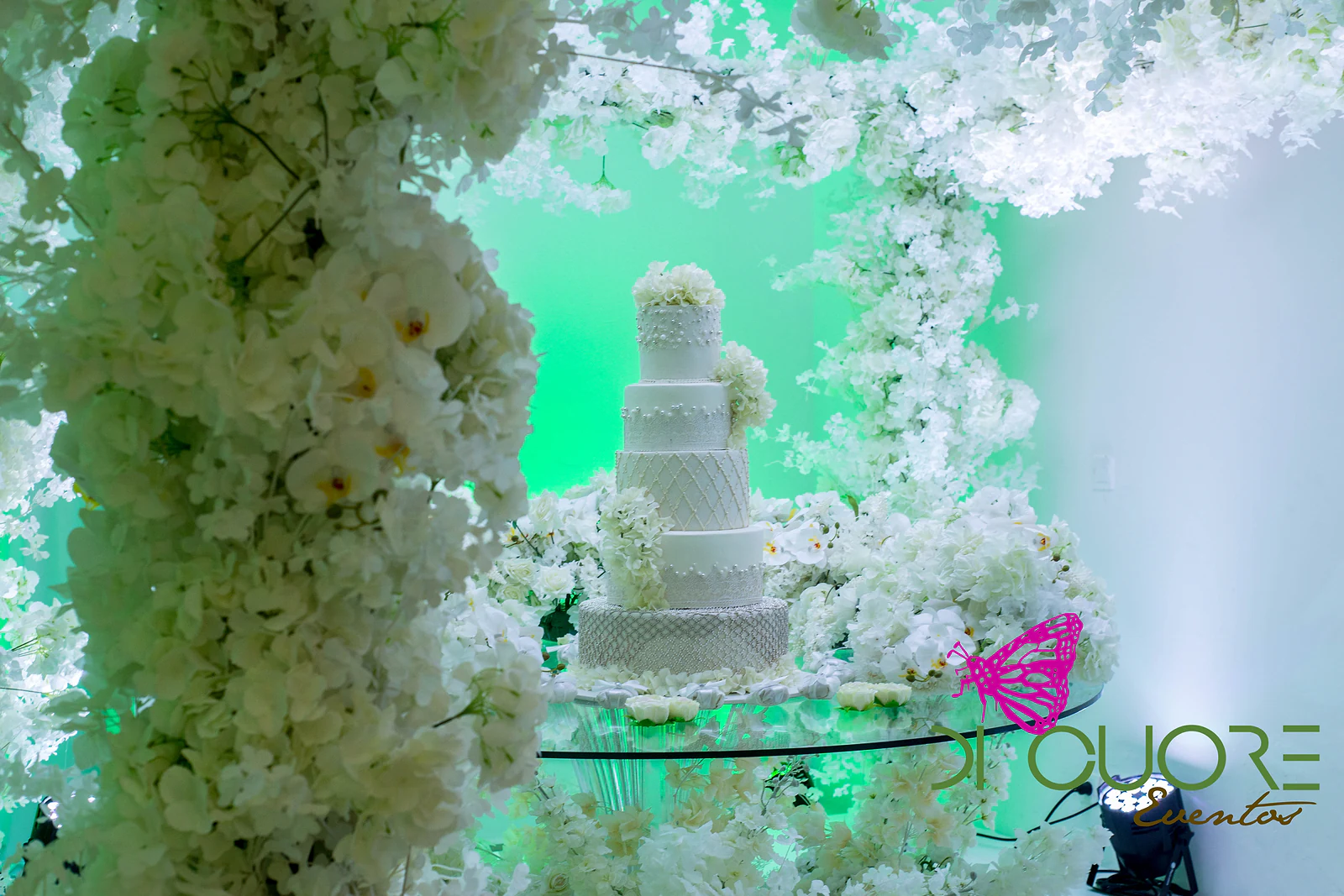 Bolo de cinco andares branco, de pormenores rendados e hortênsias, numa mesa de vidro entre flores brancas e orquídeas