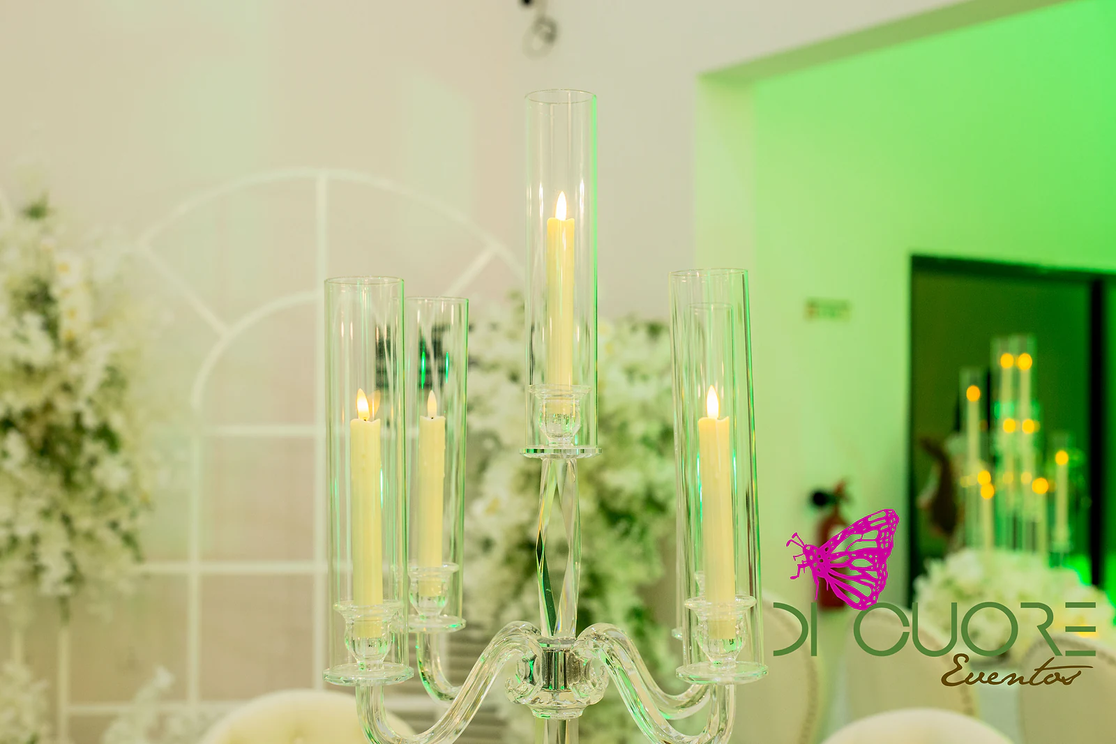 Candelabro de vidro com cinco velas em cilindros transparentes, diante de um arco branco e de flores brancas